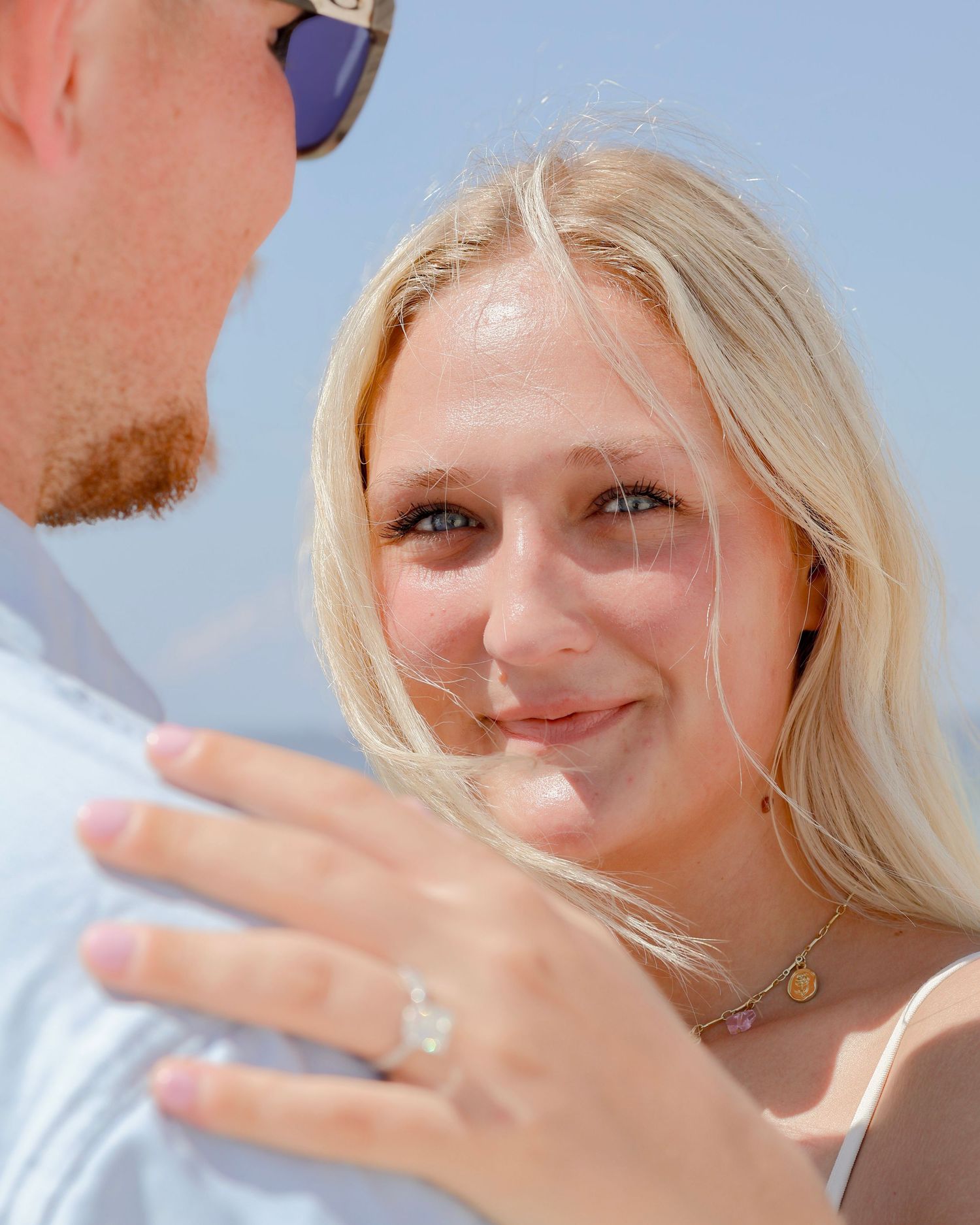 A close-up shot shows a smiling person with blonde hair in sunlight during an outdoor engagement photo.