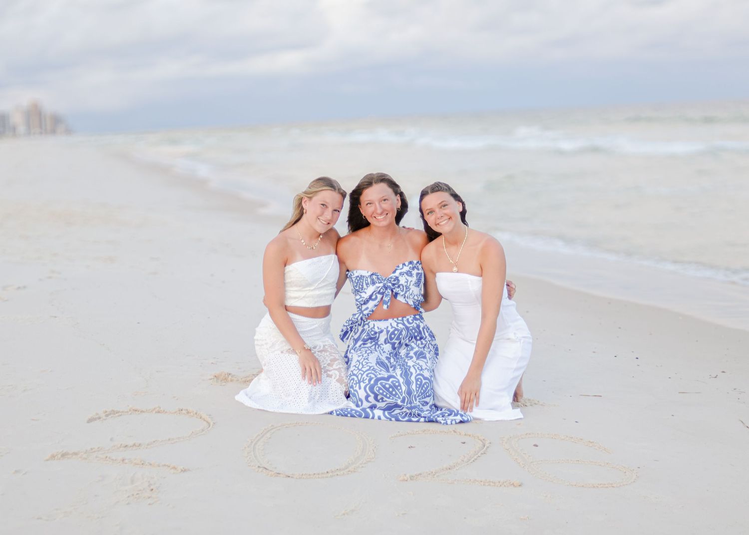 Three figures in white and blue dresses pose together on a misty beach at sunset.