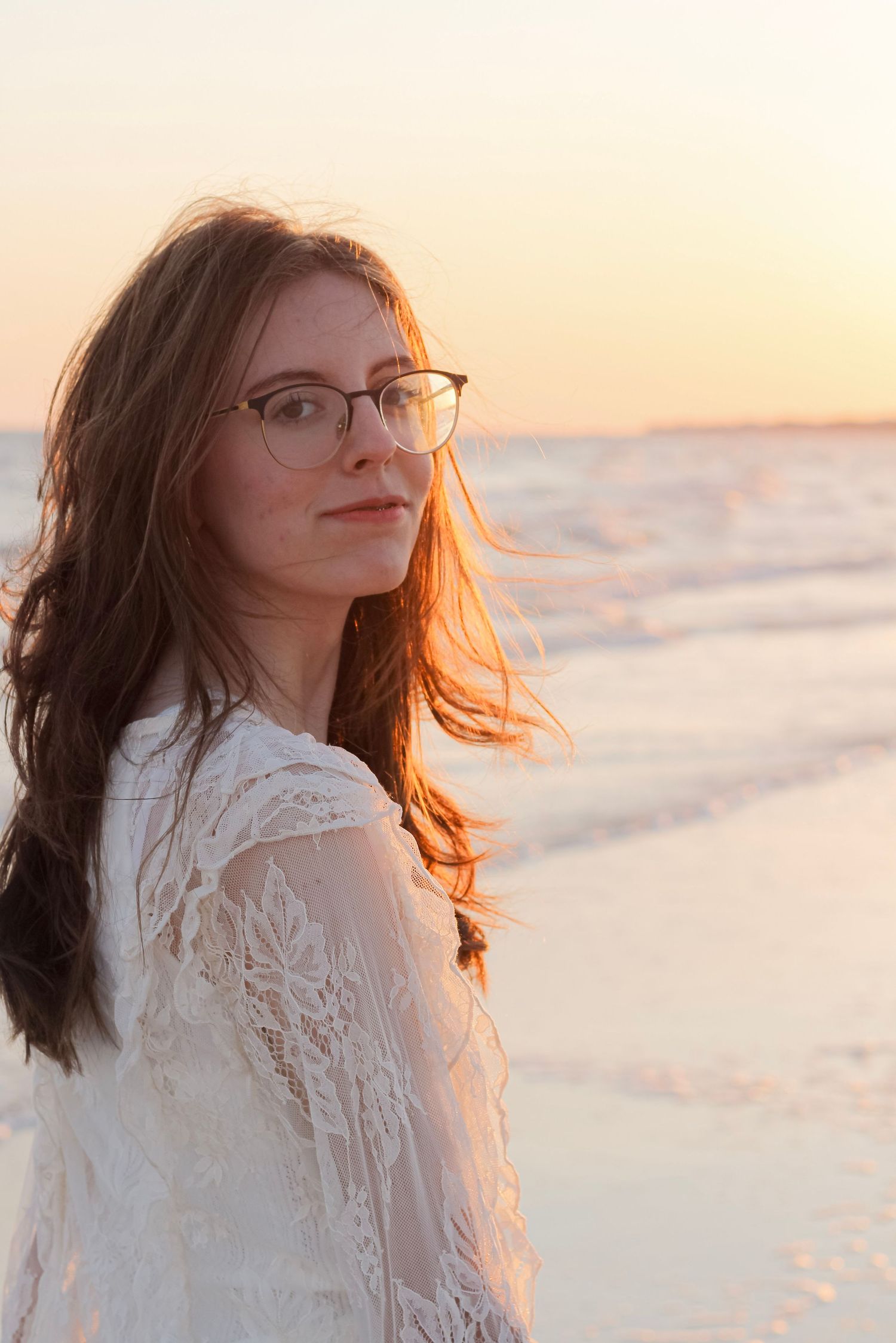 Woman in white lace dress and glasses stands by beach at sunset with hair blowing in golden light.