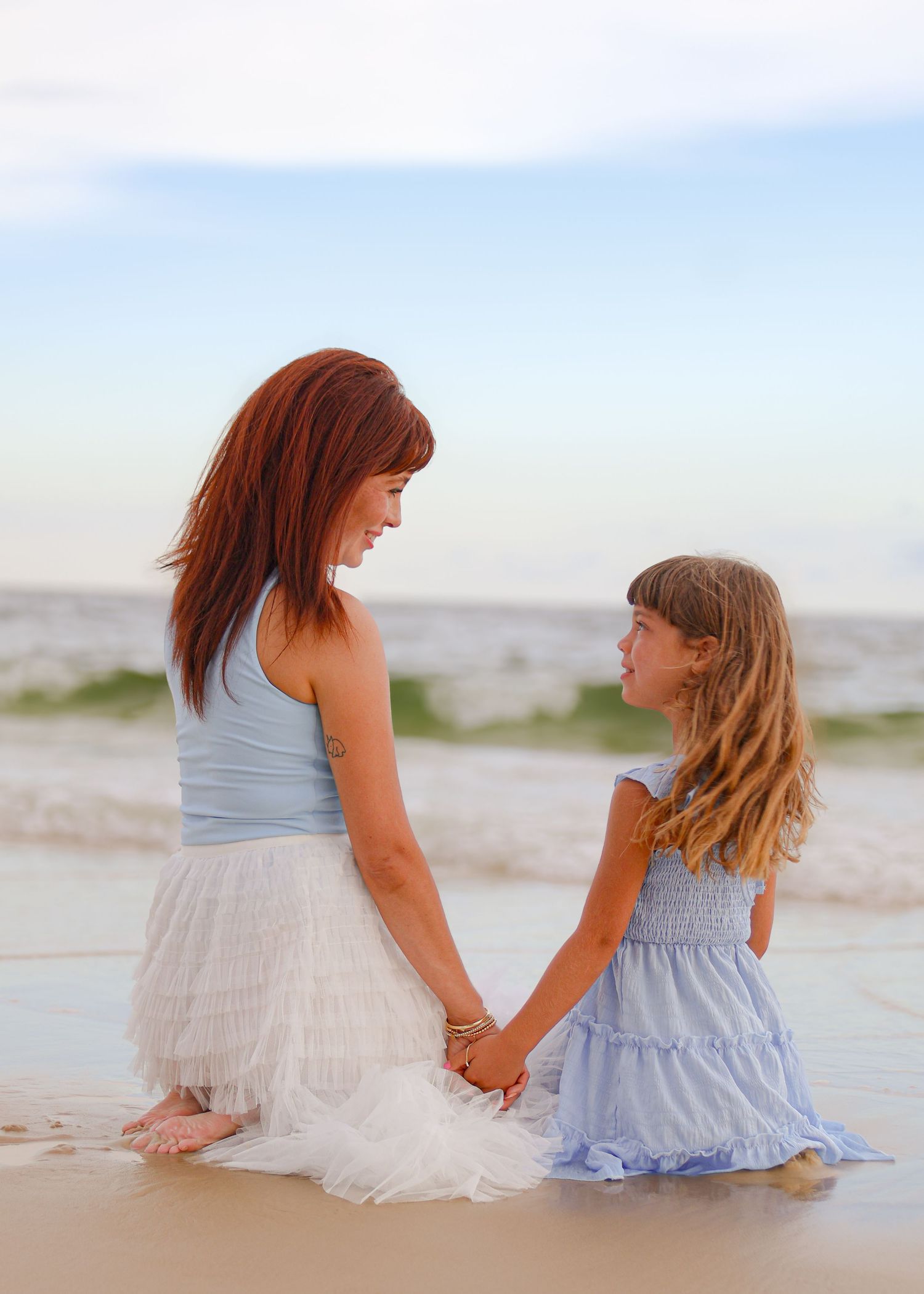 Two figures in coordinating blue and white dresses sit together on a sunny beach gazing at the ocean waves.