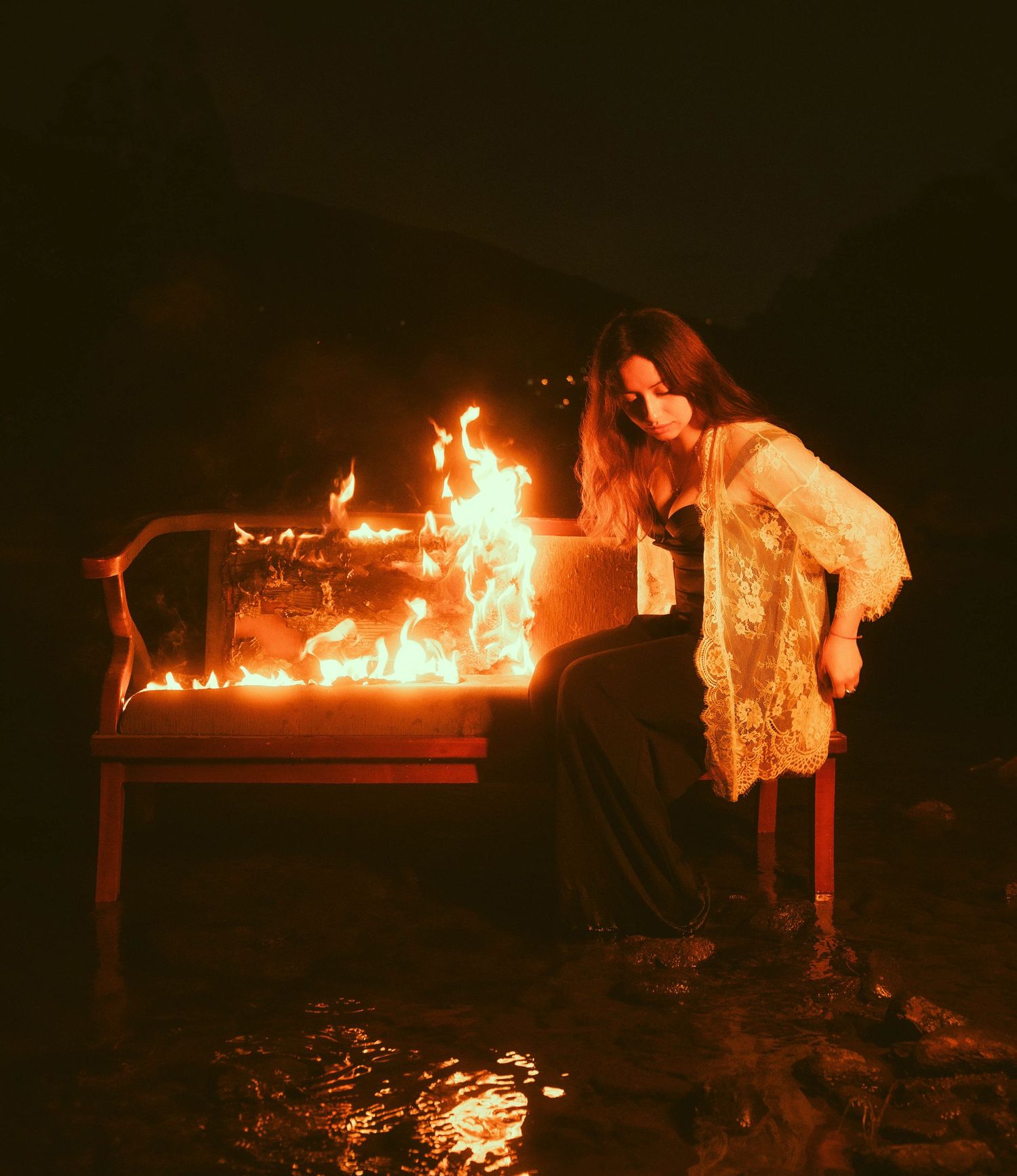 A person in lace sits beside a burning piano creating an artistic nighttime scene.