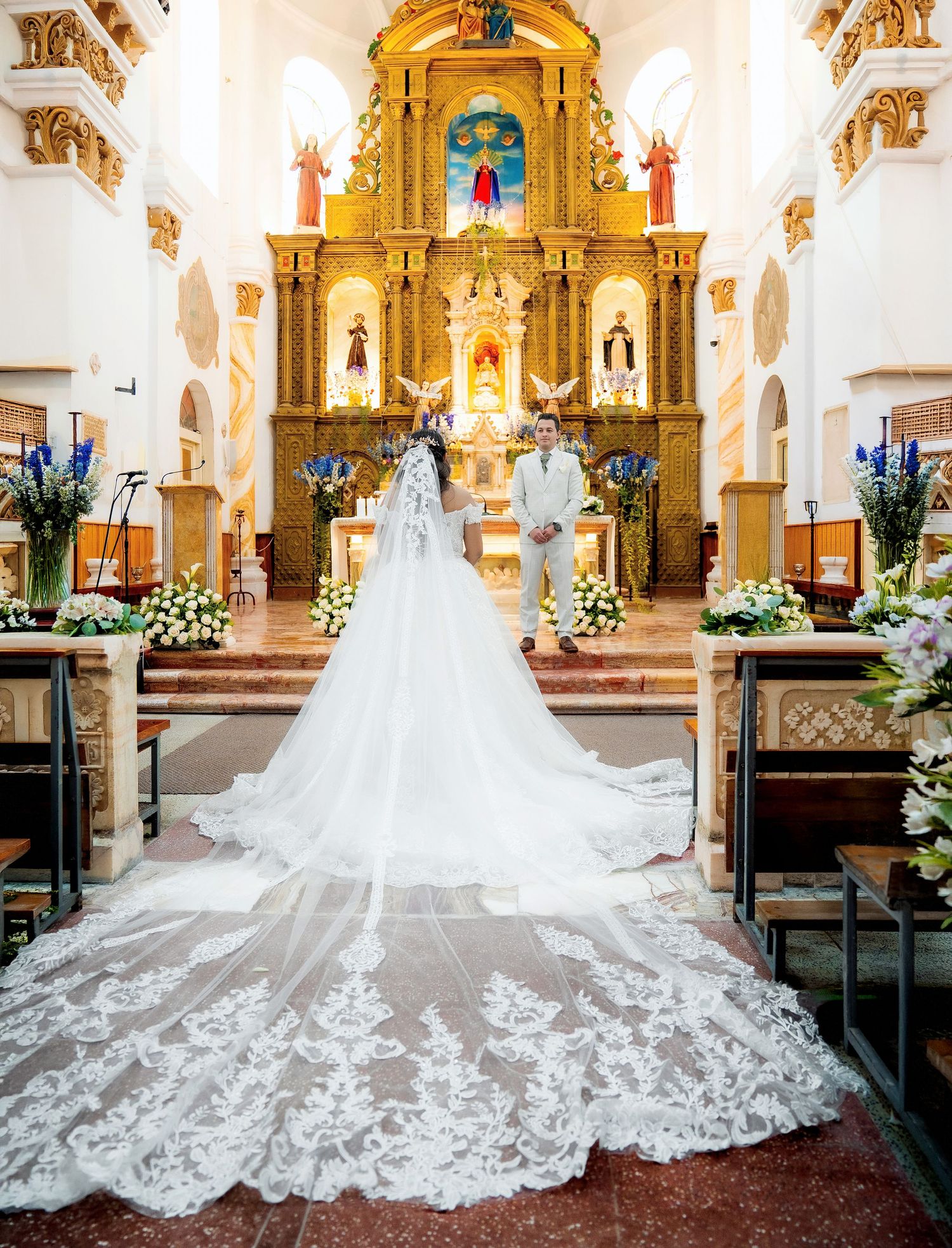 A bride in a flowing white wedding dress with a dramatic lace train stands in an ornate church with golden altar details.