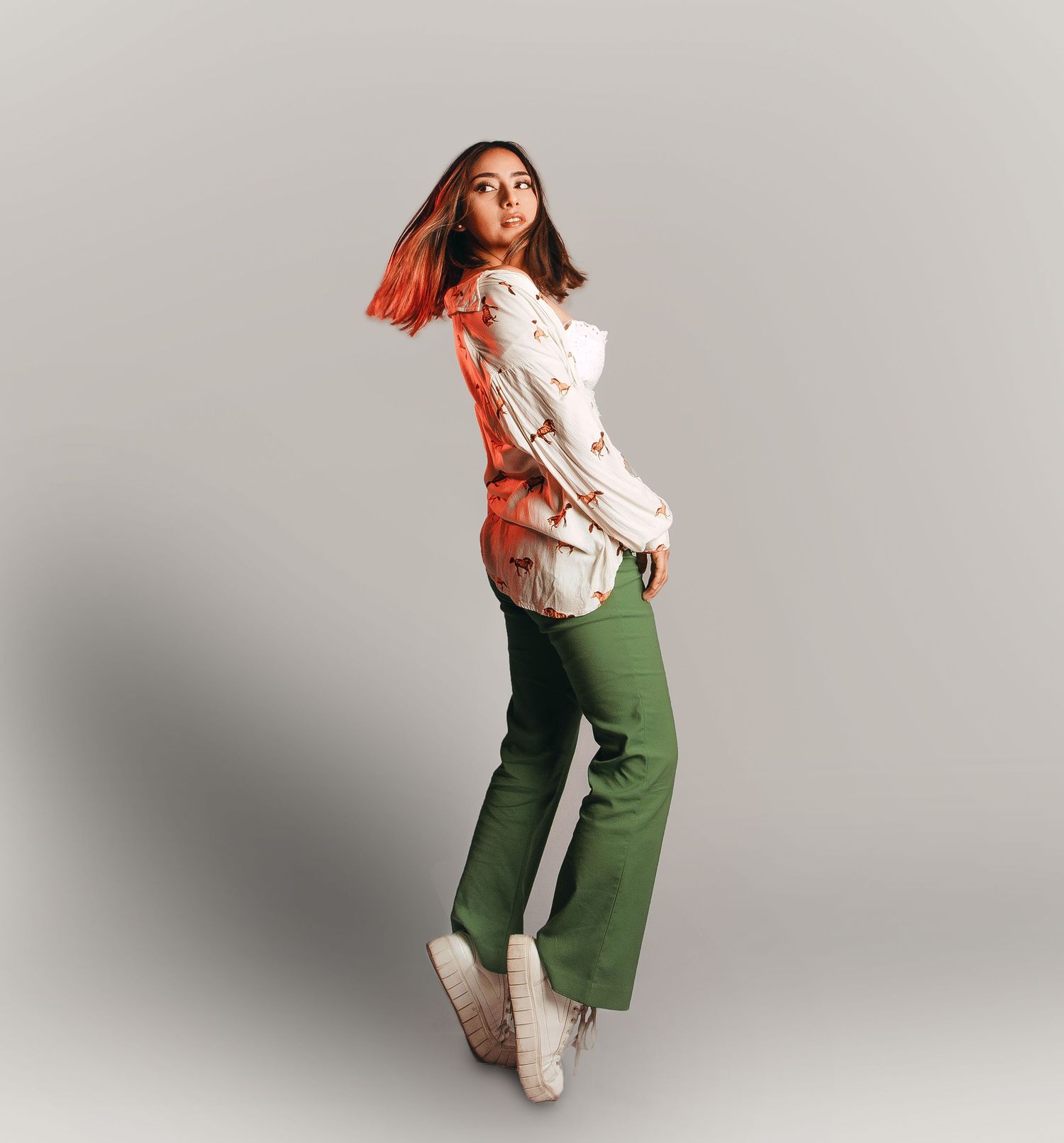 Fashion model in white top and green pants poses dynamically against grey background.