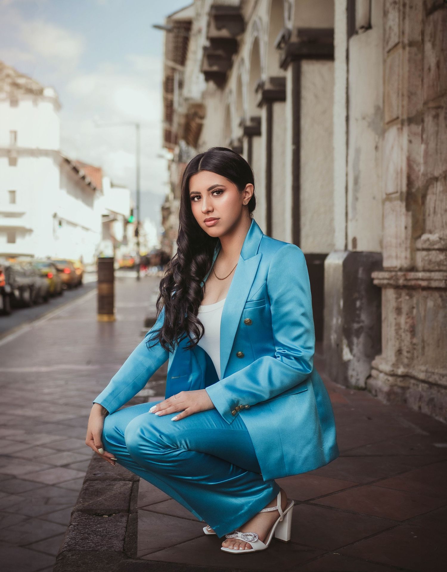 Figure in bright turquoise suit crouches on city sidewalk beside historic buildings.
