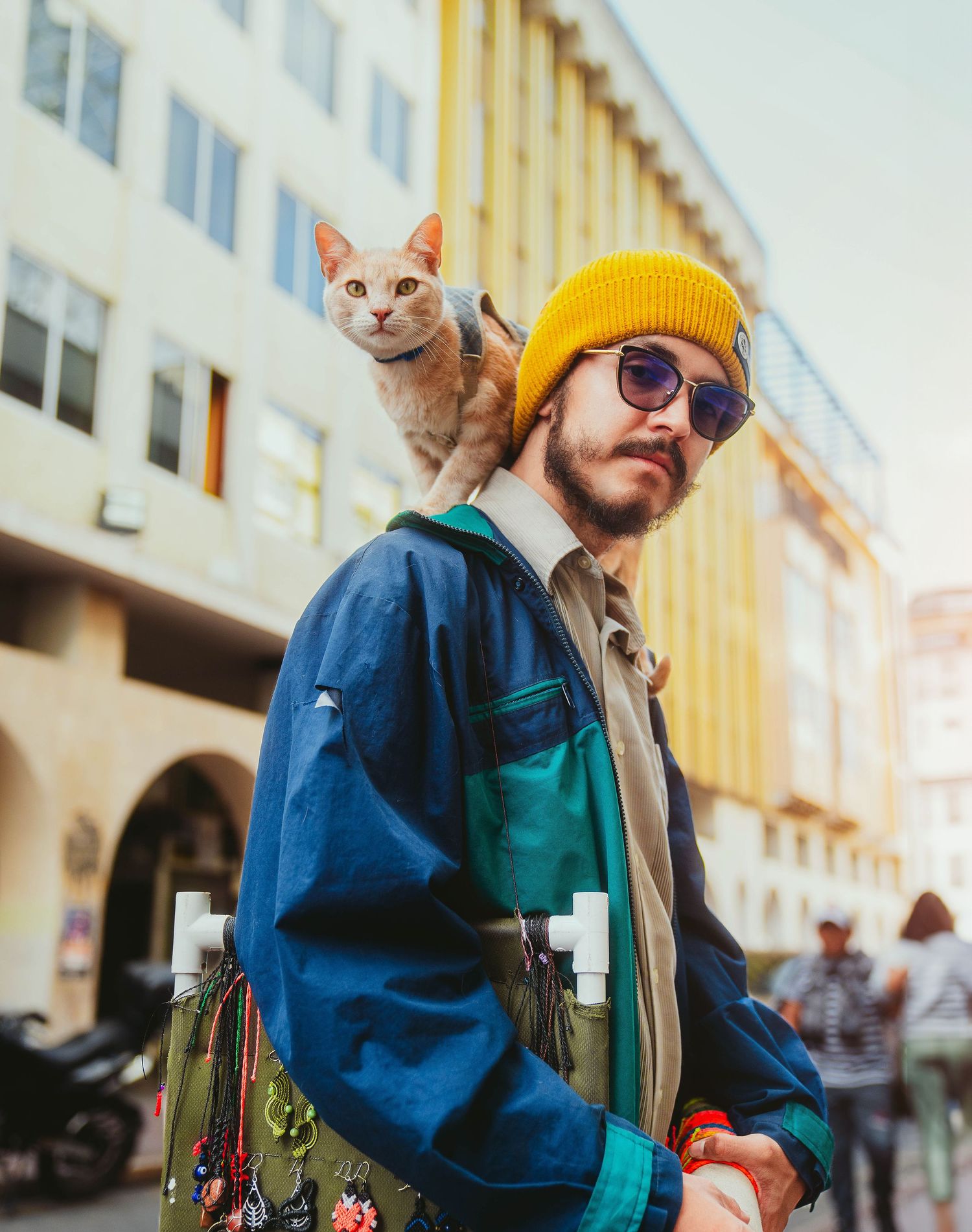 Stylish person in yellow beanie and blue jacket with orange cat on shoulders against urban backdrop.