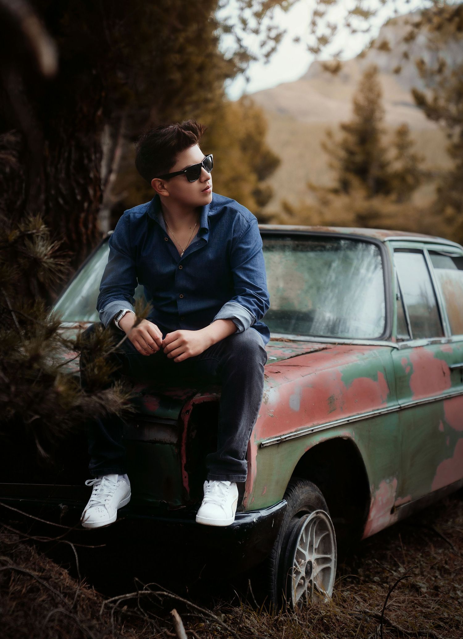 Person sits casually on vintage car wearing sunglasses and blue shirt against mountain backdrop.