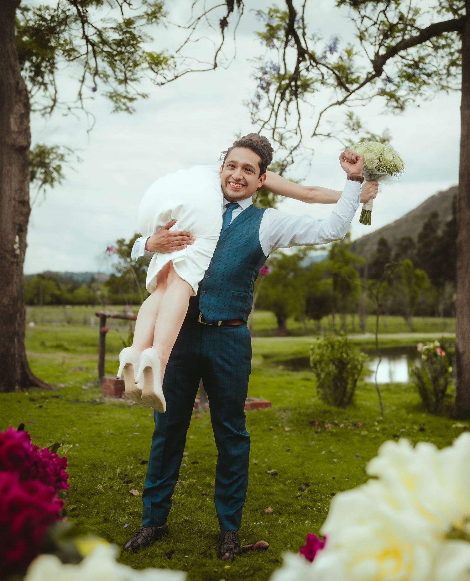 A joyful groom in blue vest holds wedding accessories while posing in a garden setting.
