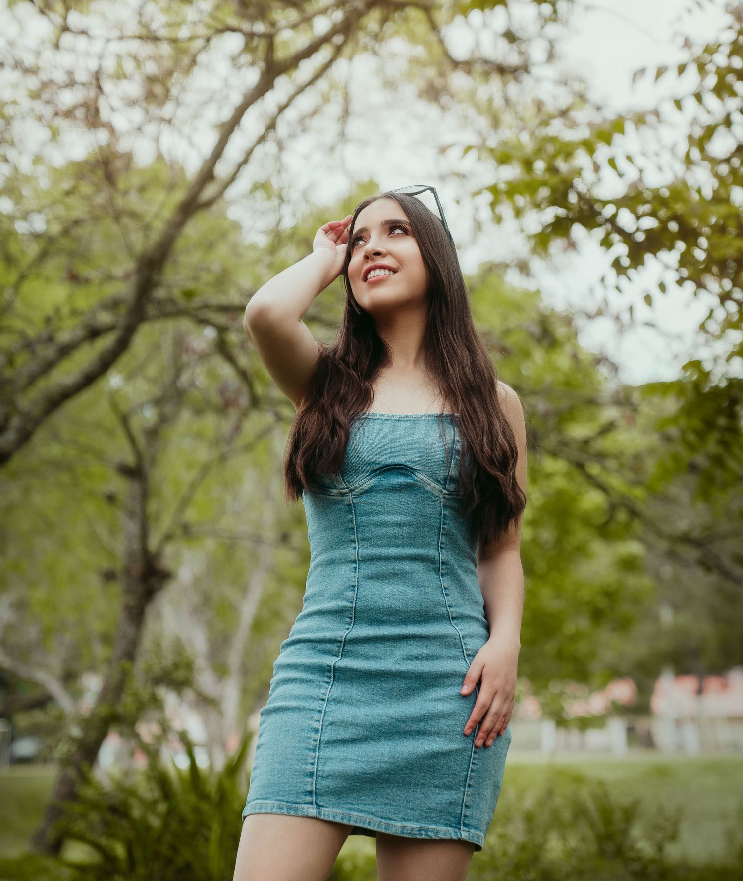 Woman in teal denim dress poses outdoors among green trees on sunny day.