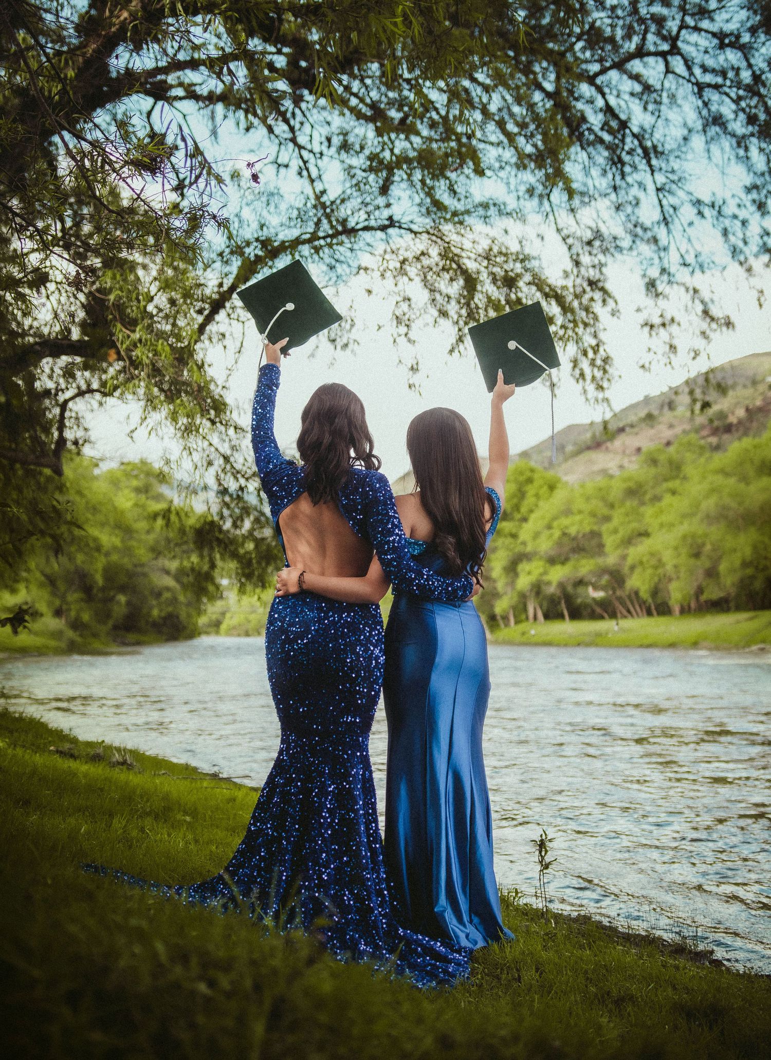 Two graduates in sparkly blue dresses hold up caps by river during sunset celebration.