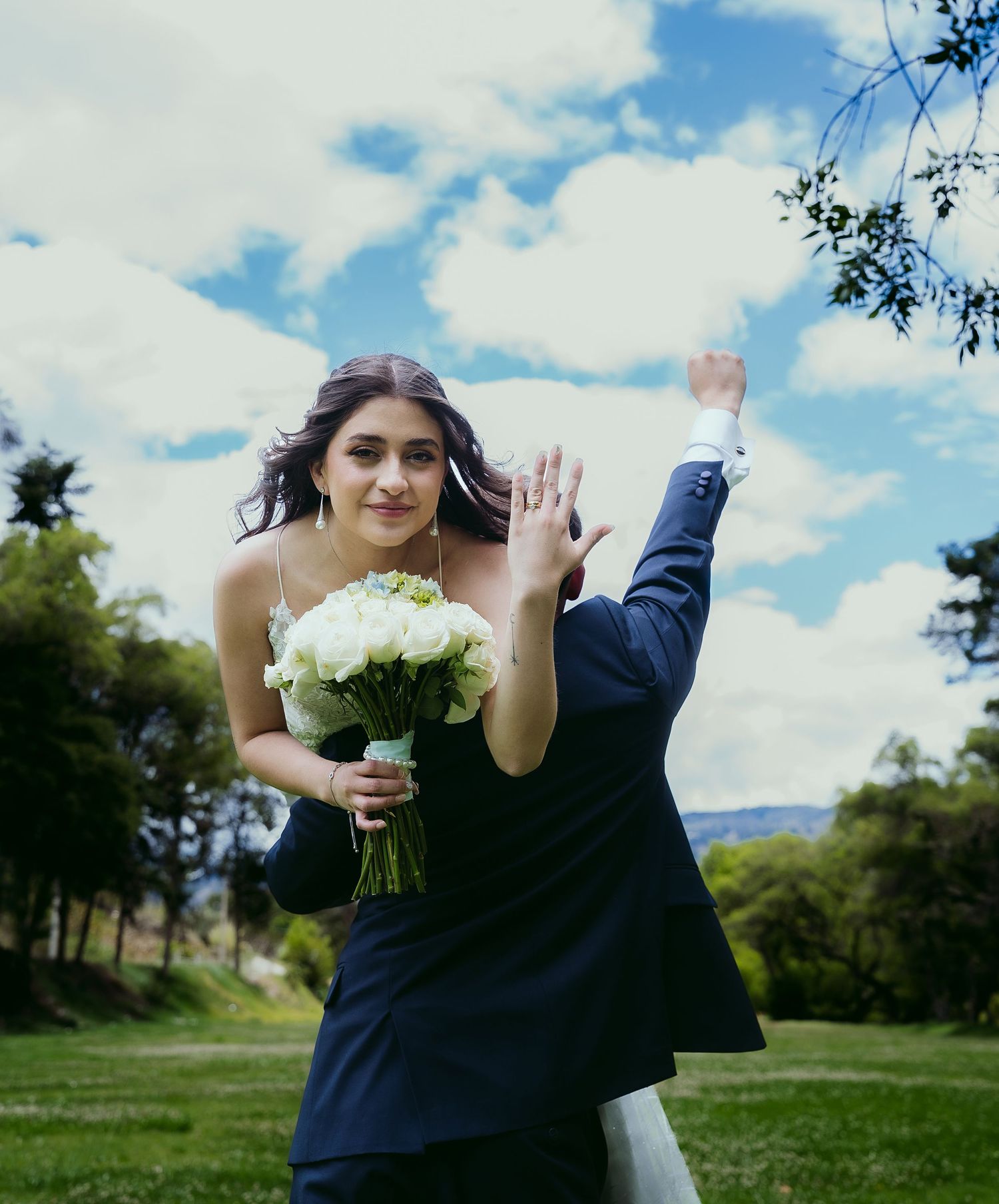 A couple embraces outdoors in a romantic pose against a bright blue sky with white clouds and green landscape.