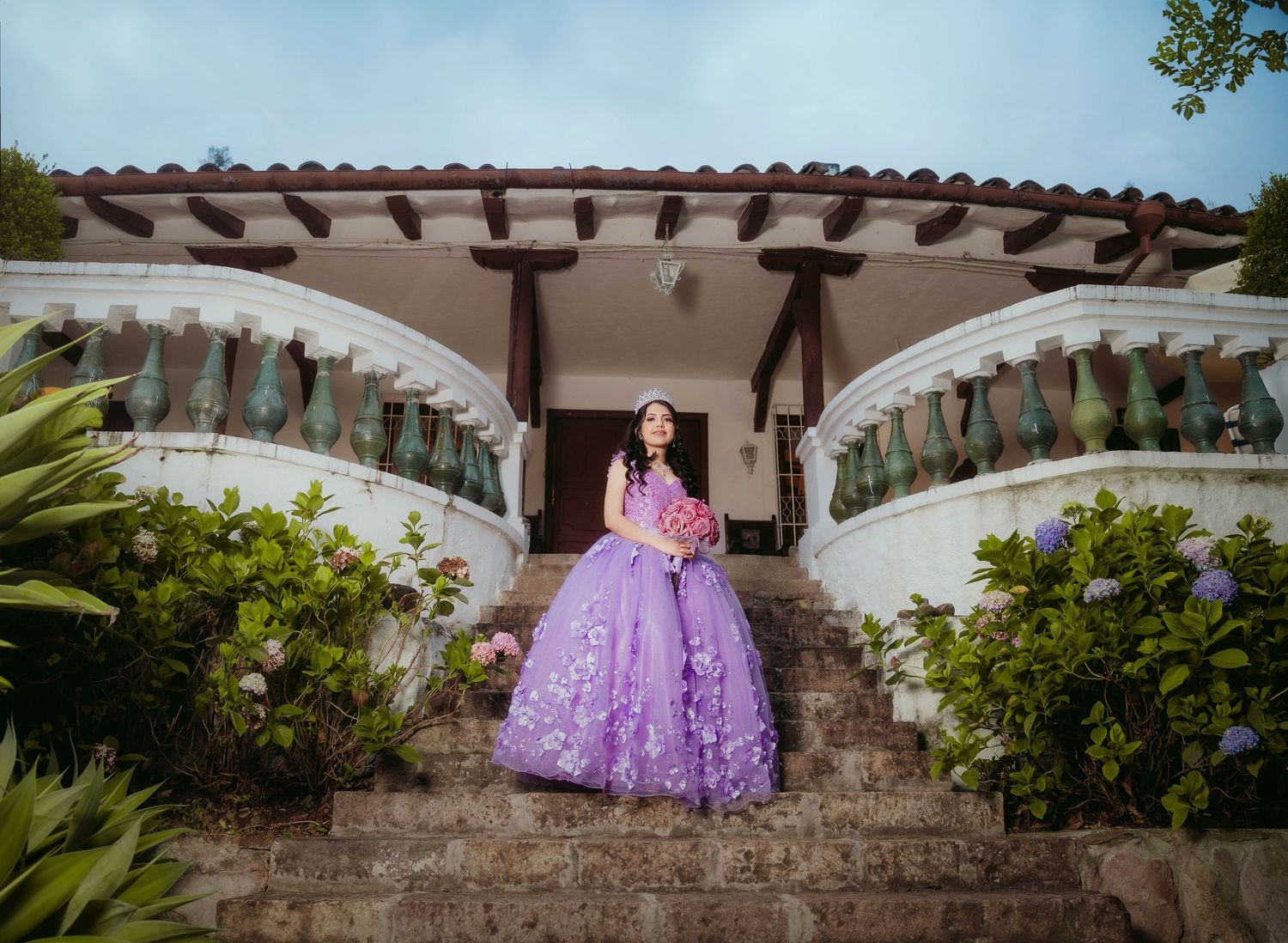 A purple quinceañera dress sparkles on the steps of an elegant Spanish-style villa with stone balustrades and greenery.