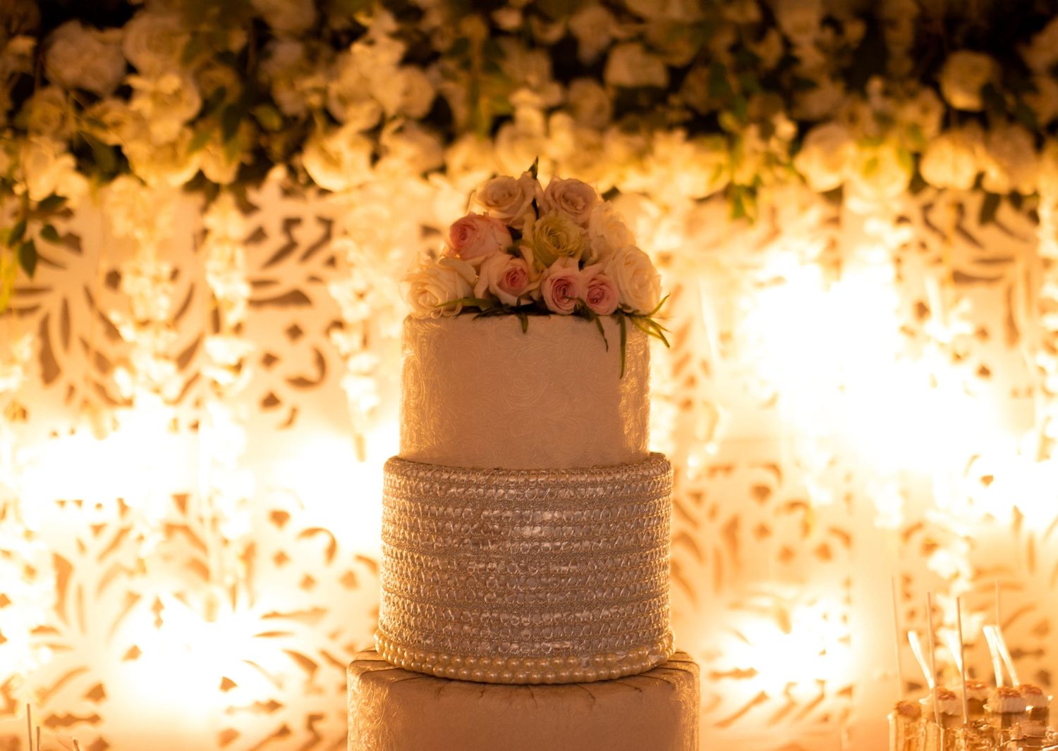 A lit wedding cake on display with decorative white details and lighting effects in the background.