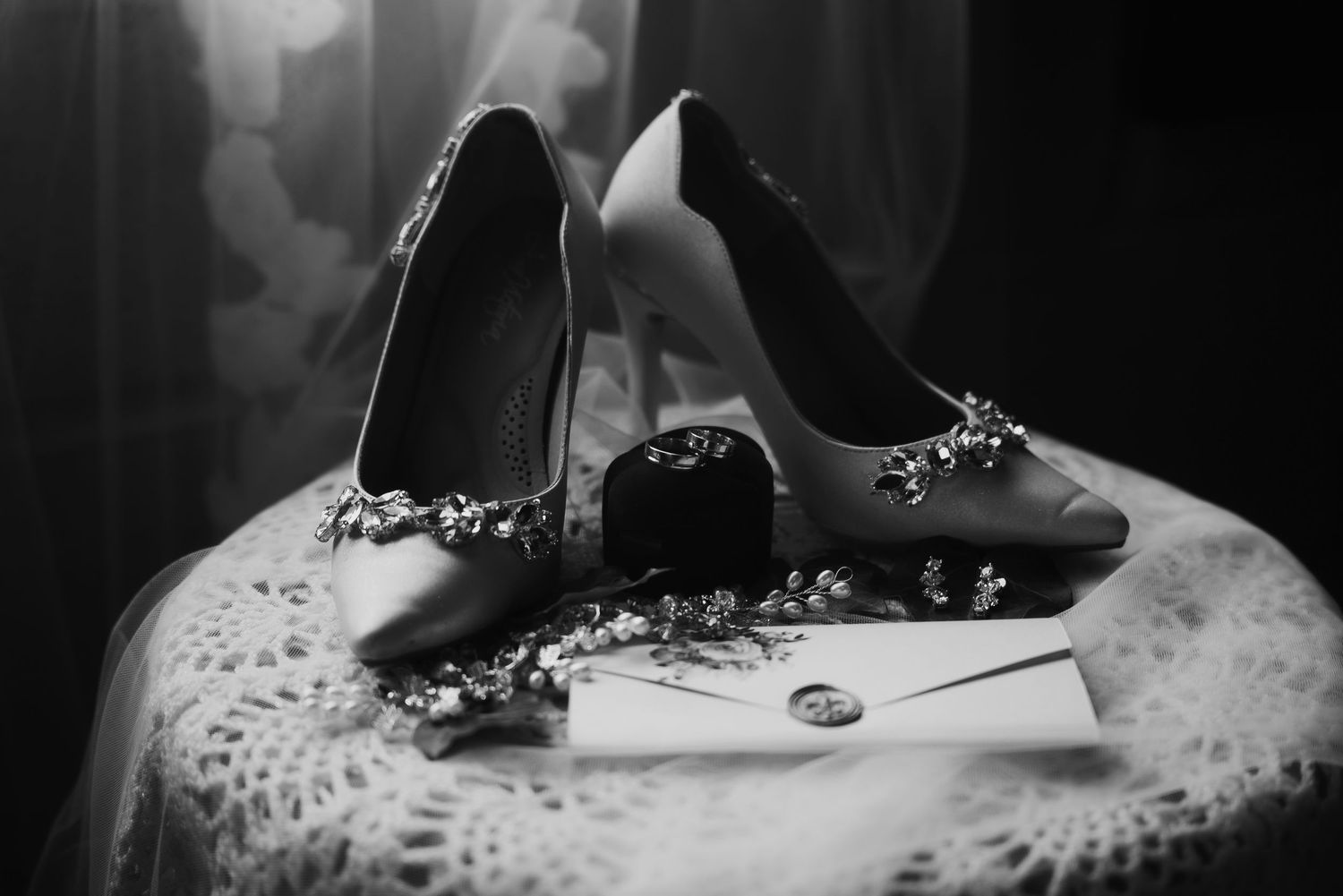 Black and white photo of elegant bridal heels with floral details, a ring, and an envelope on a lace table.