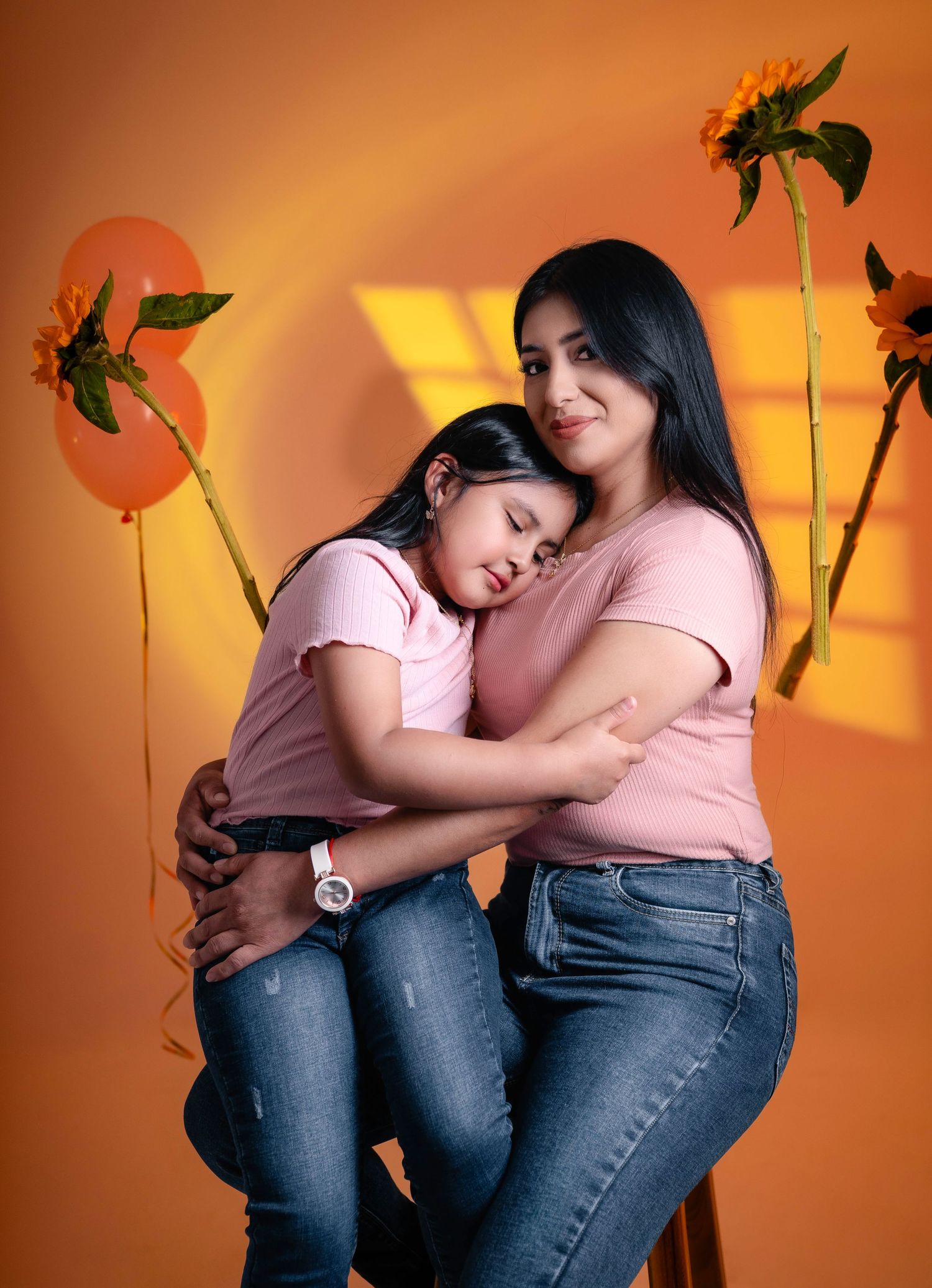 Two people in pink tops and jeans share a warm embrace against an orange backdrop with decorative sunflowers.
