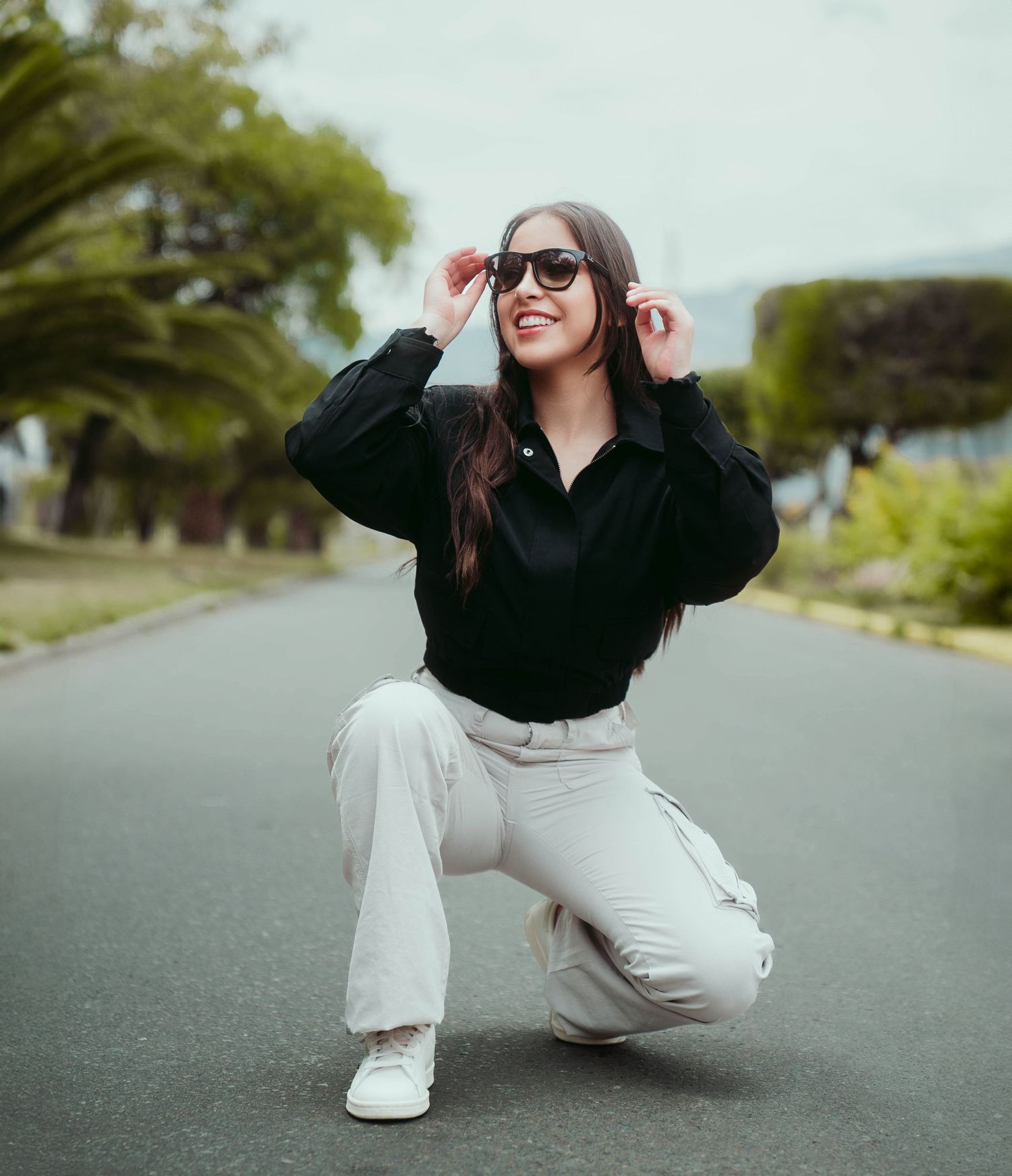 Person in black sweater and white pants crouches on suburban street wearing sunglasses.