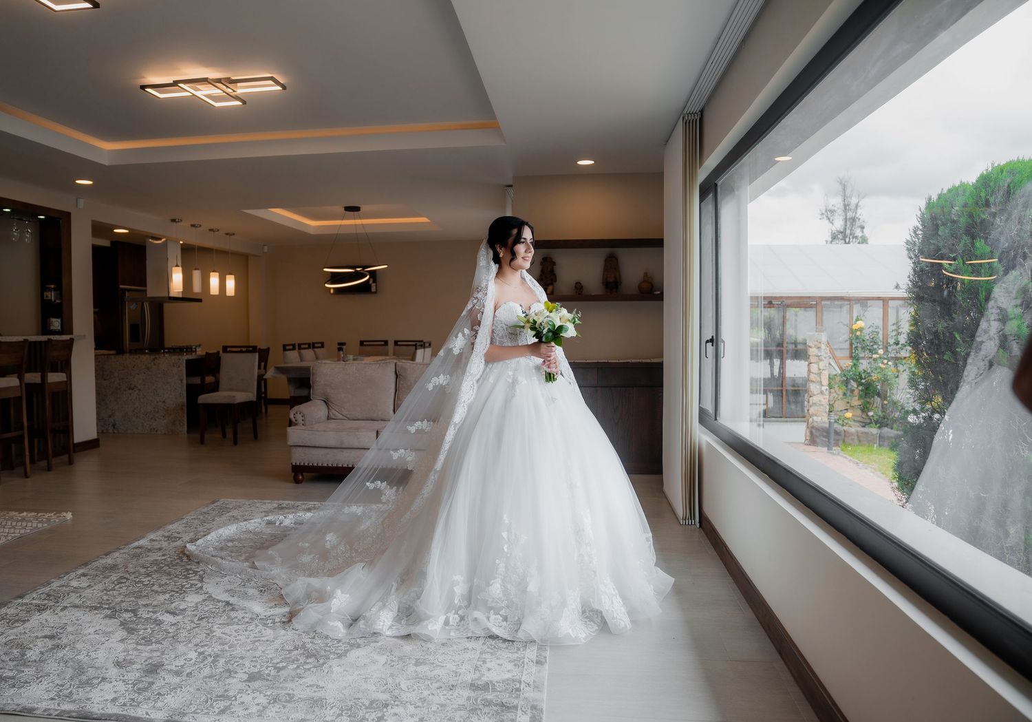 Bride in elegant white ball gown with long veil standing by large window in modern wedding venue.
