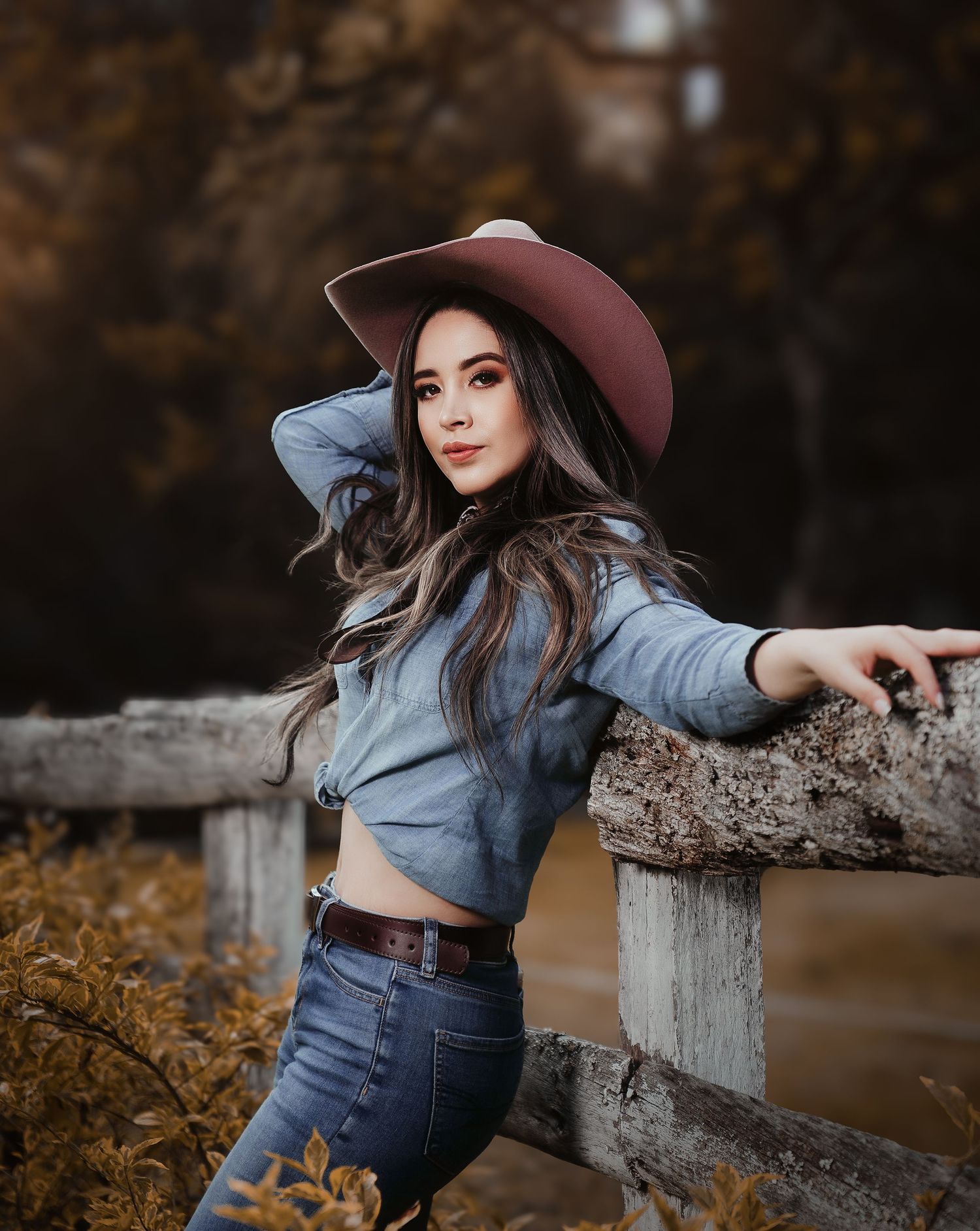 A stylish woman in a wide-brimmed hat and denim outfit poses against a rustic fence during autumn.