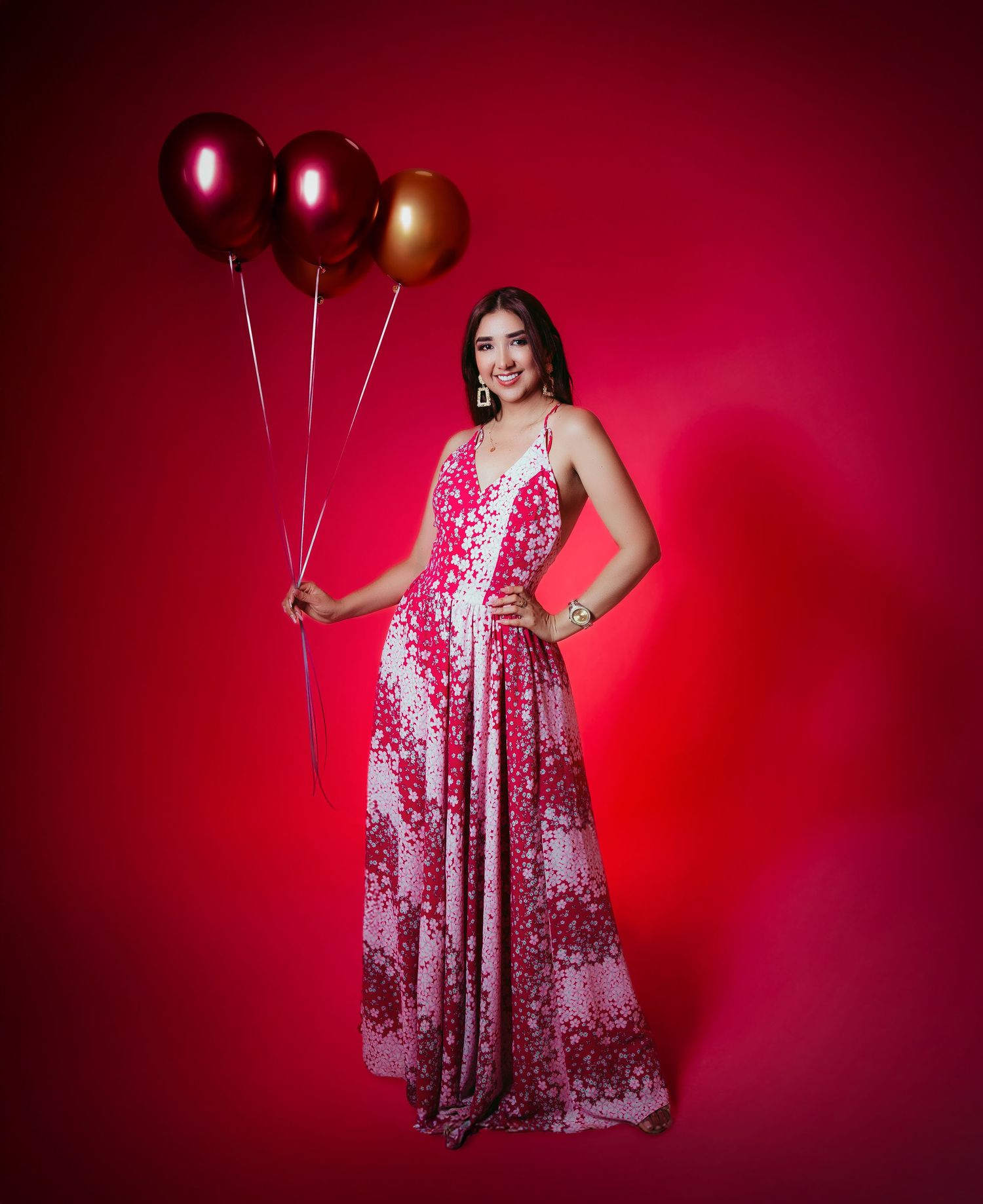 Woman in flowing pink patterned dress holds metallic balloons against red backdrop.