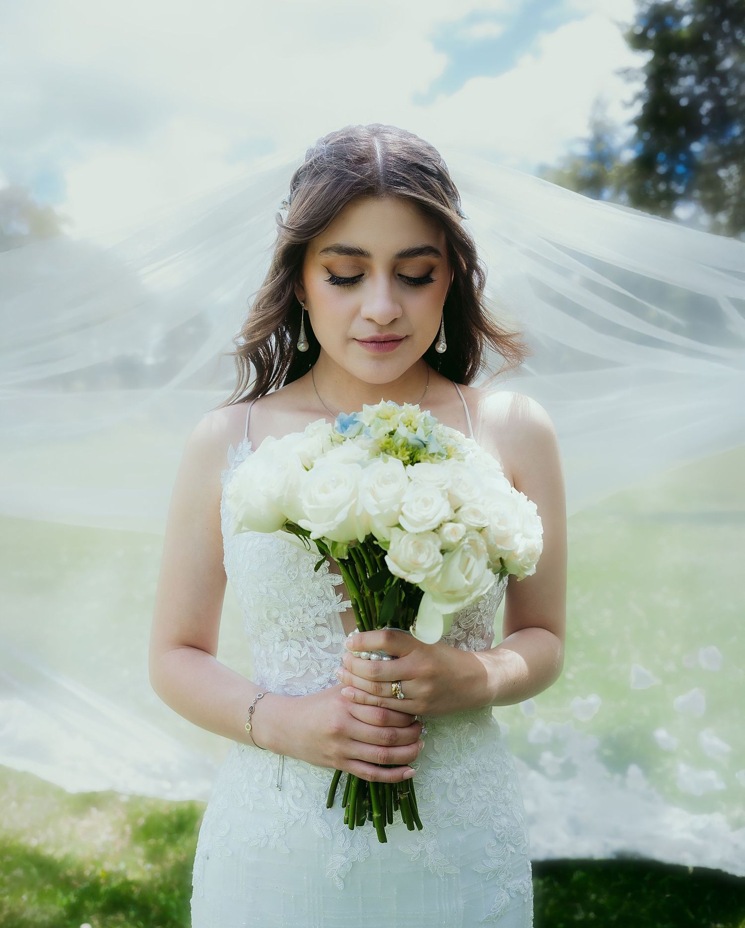 A bride in a white lace dress holds a bouquet of white flowers as her veil flows in the breeze on a sunny day.