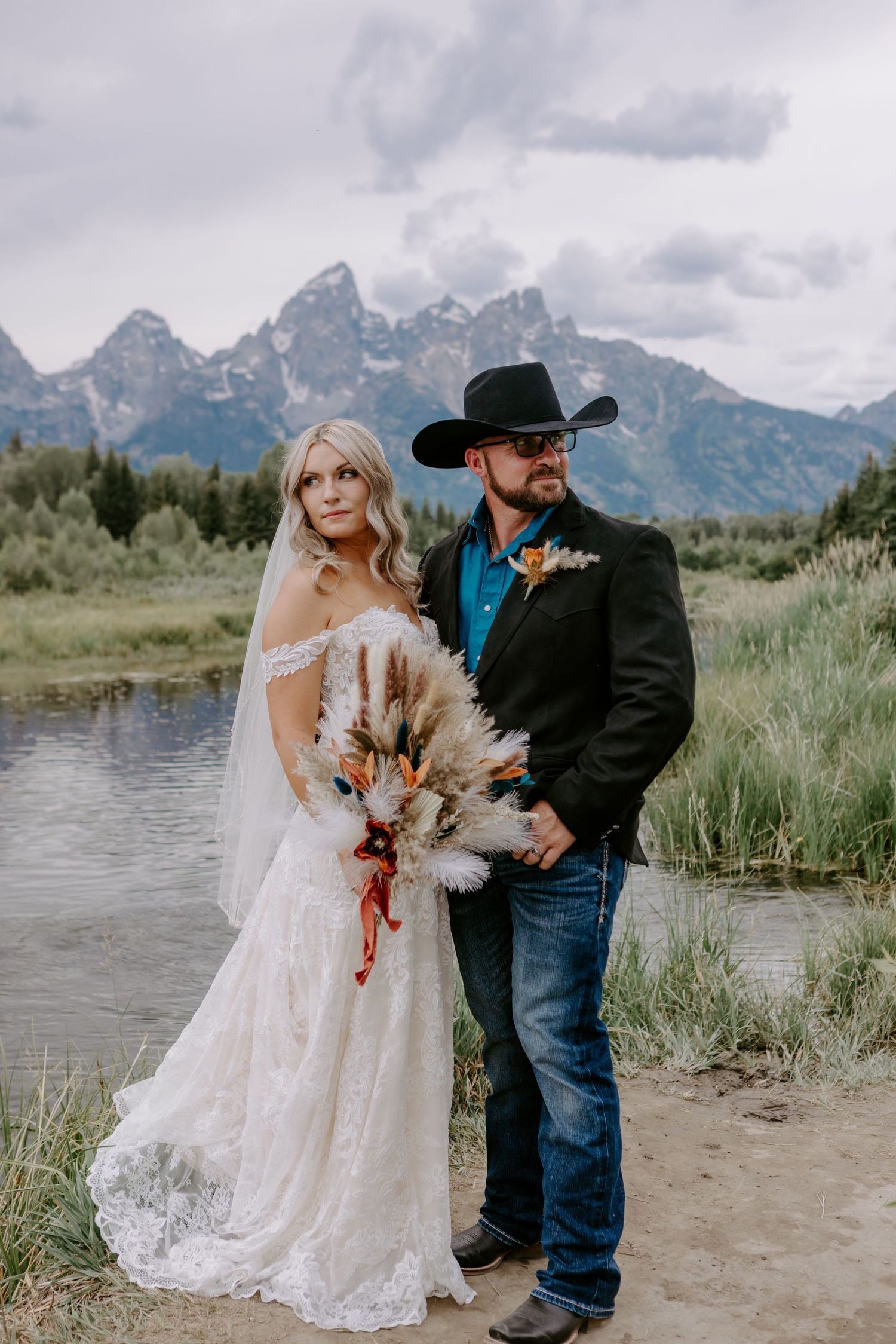 Bride and groom posing by a mountain lake in Grand Teton National Park, groom wearing cowboy hat and black jacket.