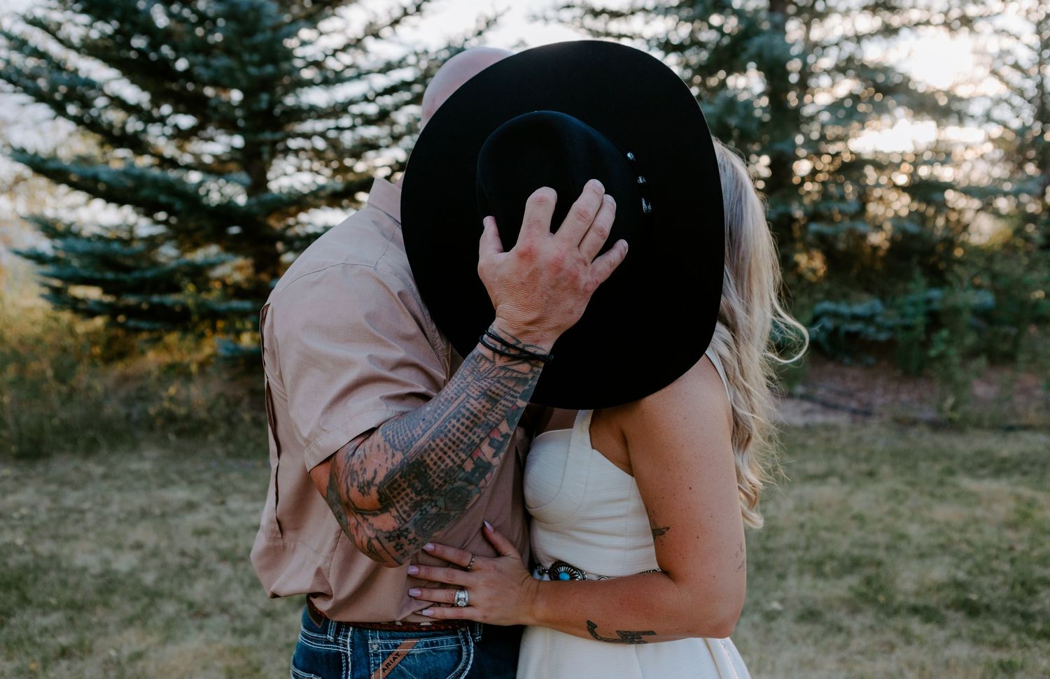 Couple sharing a kiss hidden behind a black cowboy hat, man with tattooed arm embracing woman in white dress outdoors.
