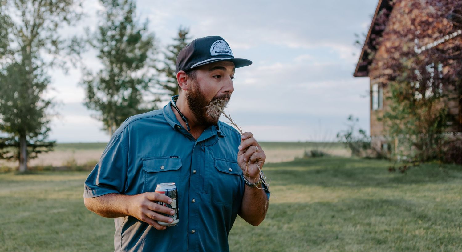 Bearded man in blue shirt and snapback hat holding a drink outdoors near a rustic building and green trees.