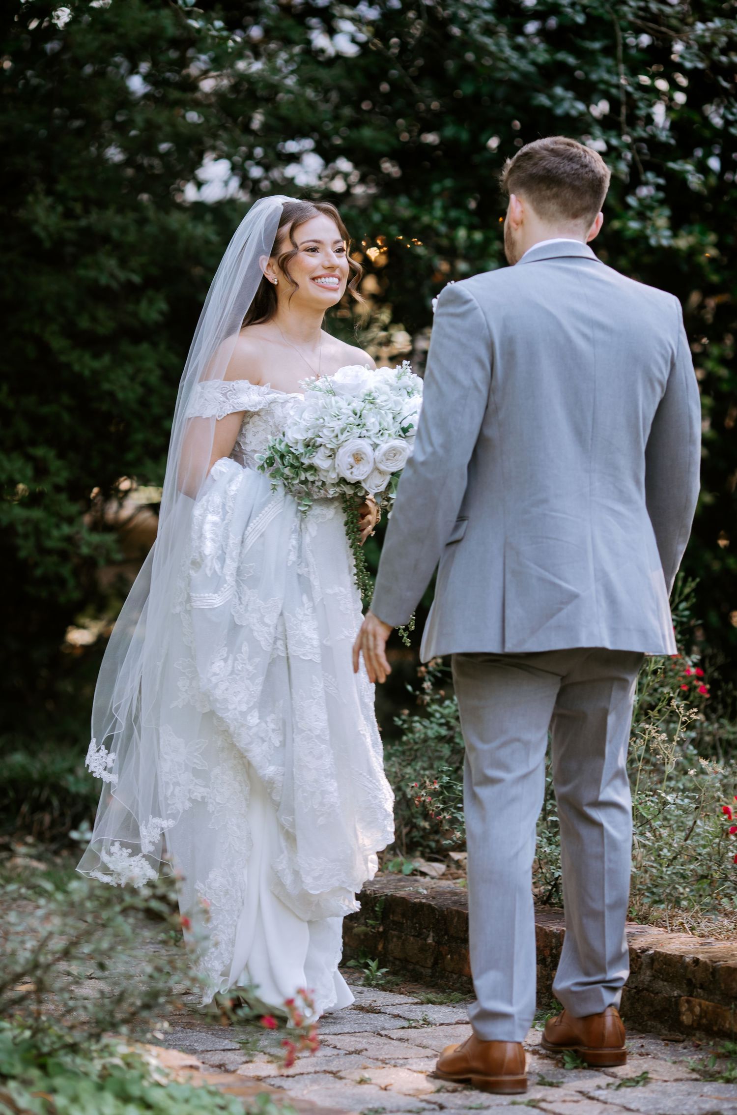 Smiling bride in off-shoulder lace gown with veil holds white bouquet facing groom in gray suit on brick path.