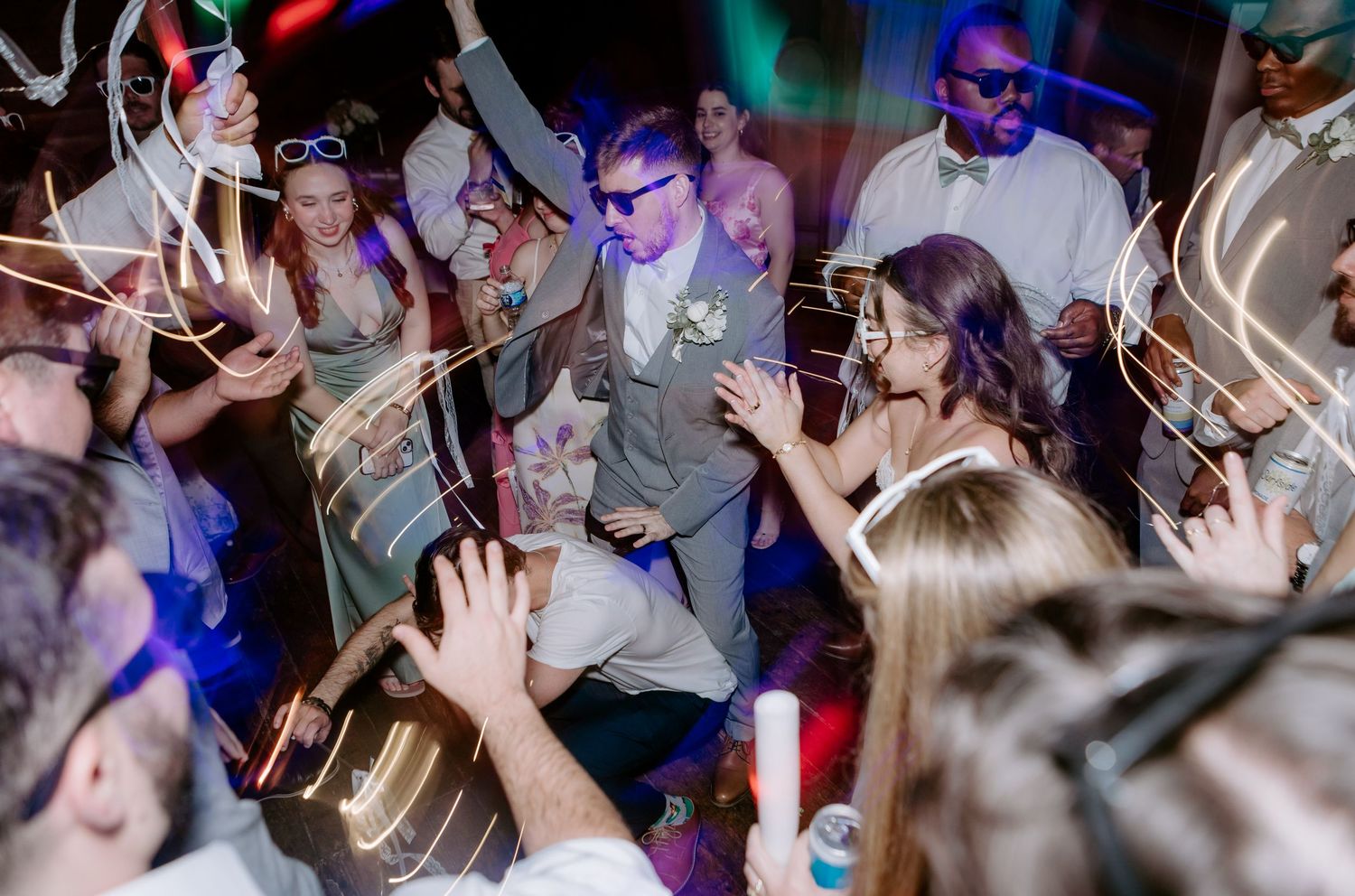 Bride and groom dance with guests under colorful lights and light trails at their wedding reception.