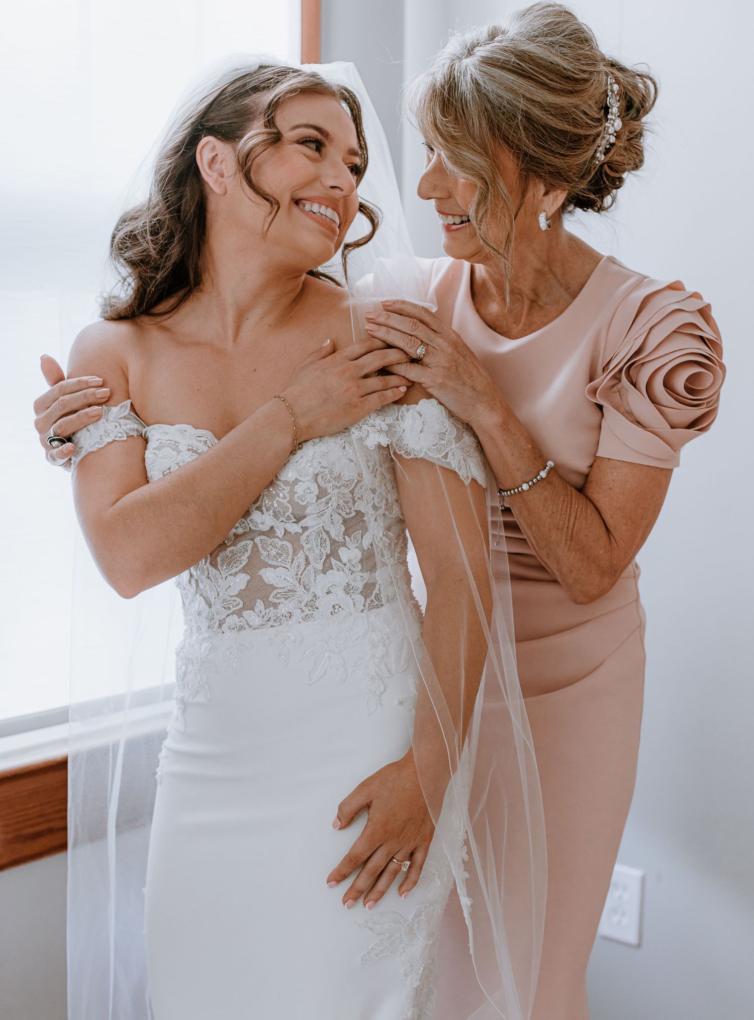Bride in white lace off-shoulder gown smiles with older woman in blush dress with rose detail sleeves during wedding preparations.