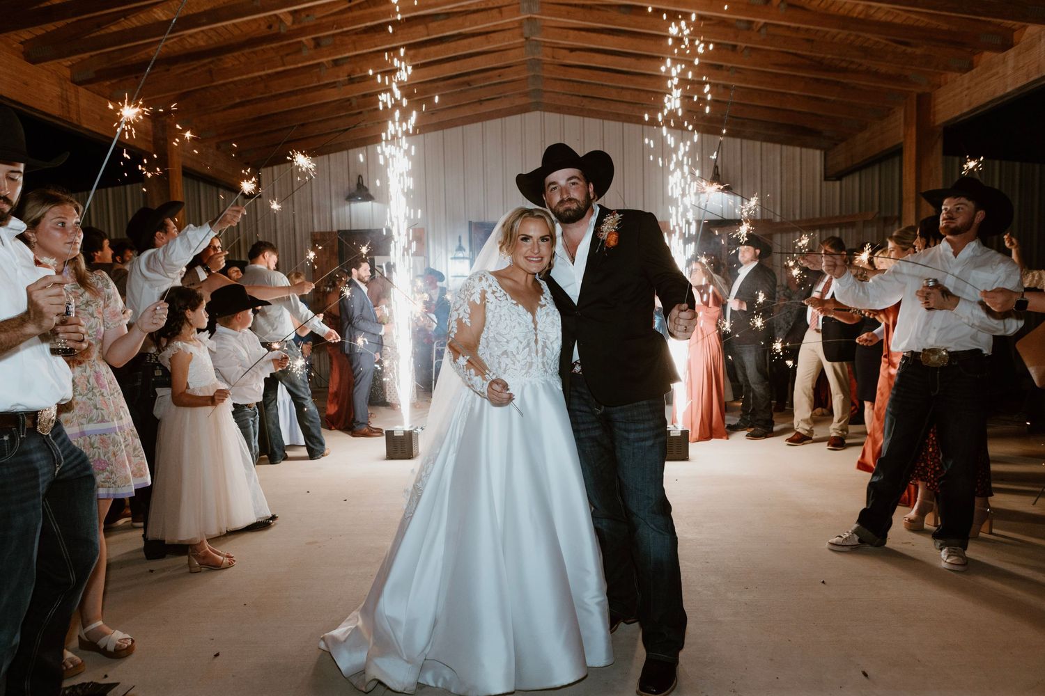 Bride and groom exit wedding reception through sparkler tunnel in rustic barn venue, wearing white gown and black cowboy hat.