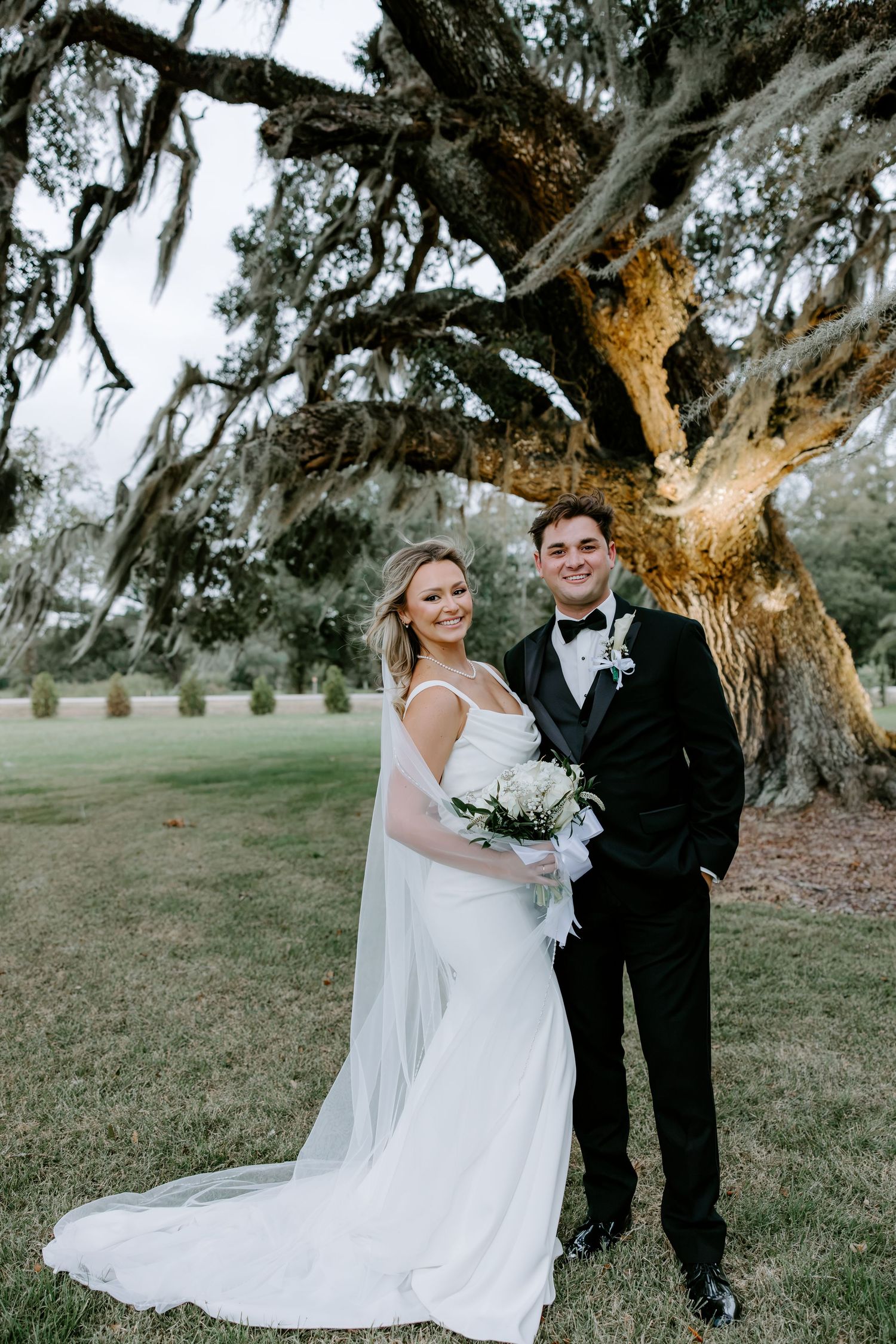 Bride in a white gown with long veil and groom in a black tuxedo smile holding a bouquet beneath a large oak draped in Spanish moss.