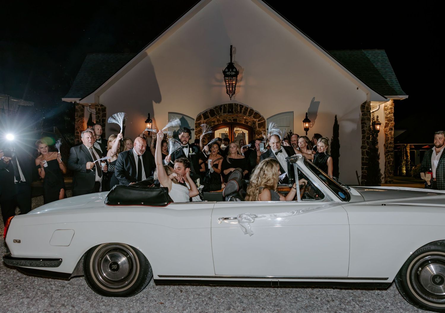 Wedding guests cheer around a classic white convertible outside a stone chapel at night during a festive send-off celebration.
