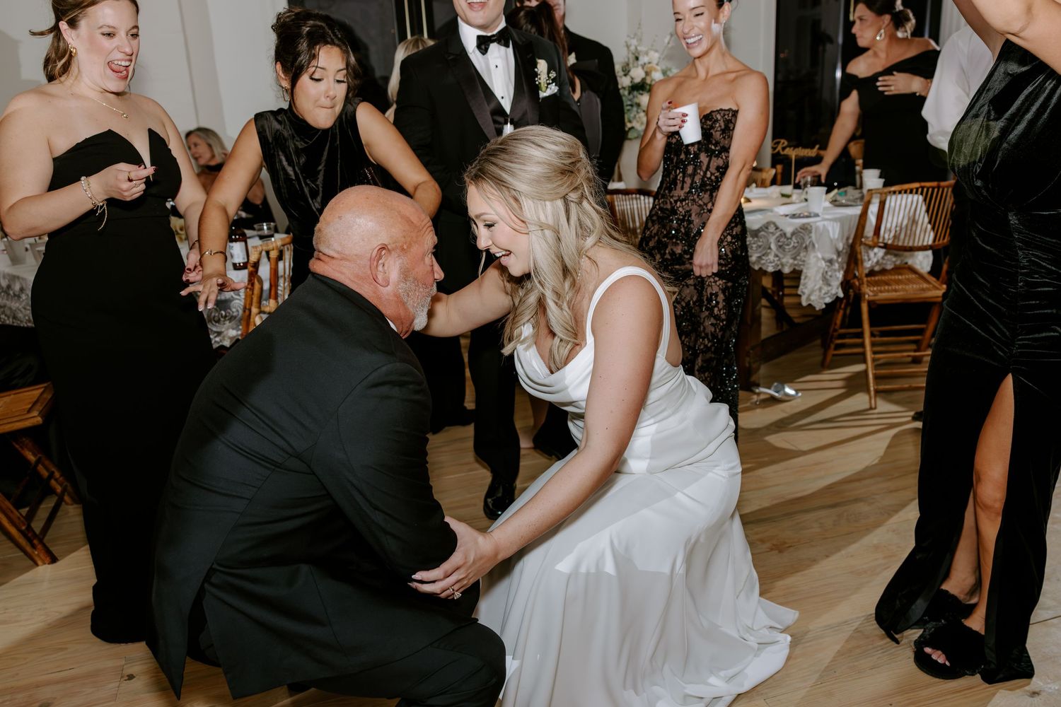 Smiling bride in a white wedding gown shares a joyful moment with a kneeling older man as guests in formal attire cheer behind them.