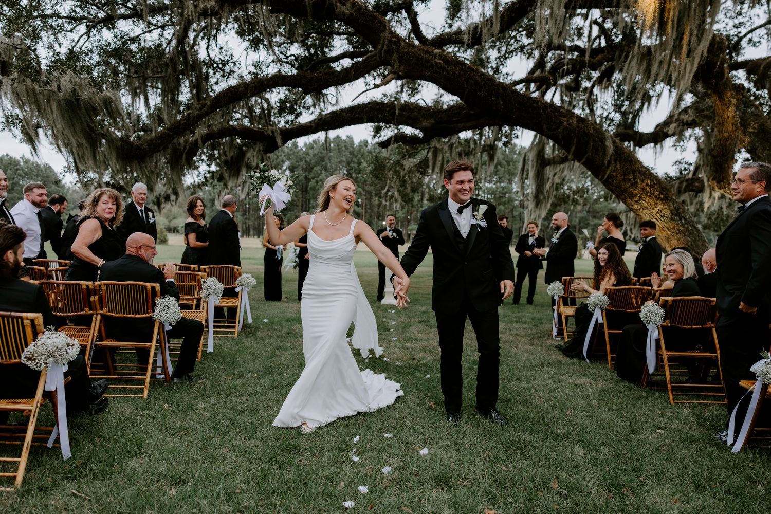 Bride and groom hold hands and walk down the aisle between guests during an outdoor wedding ceremony beneath a mossy oak tree.