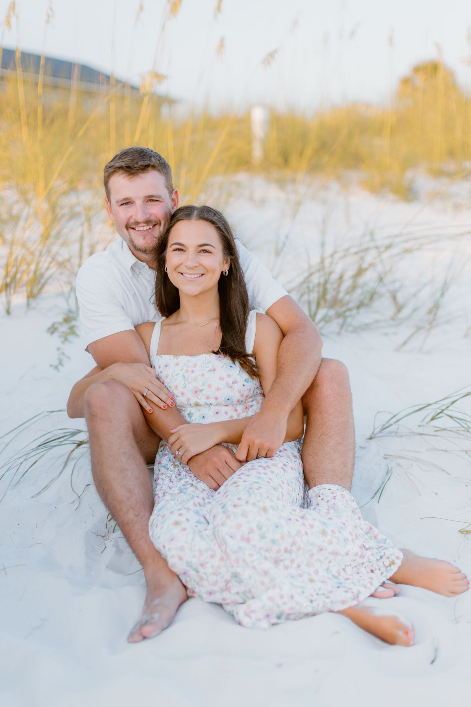 A couple sits together on white sand beach at sunset with sea oats in the background.