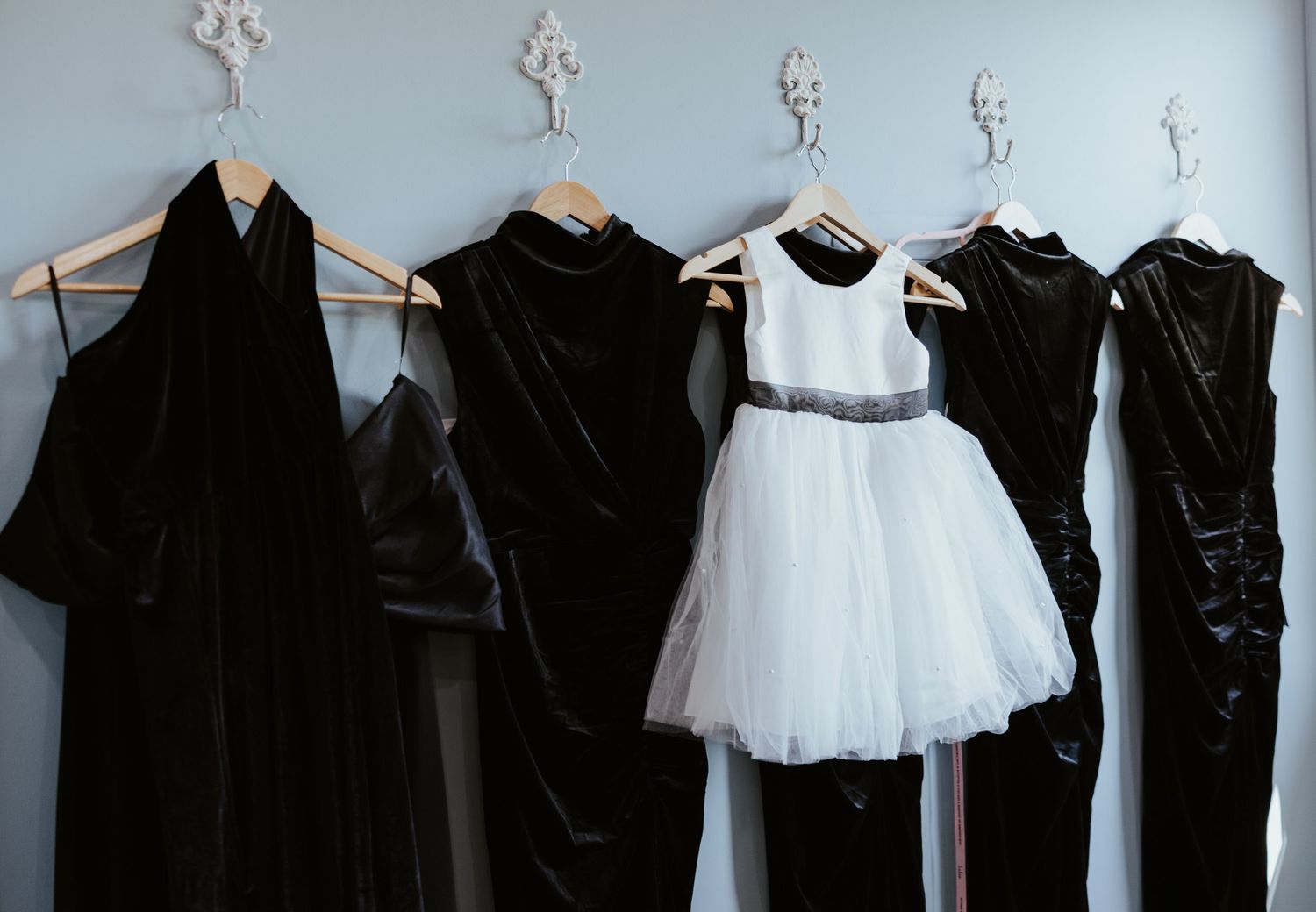 Black velvet dresses and a white tulle flower girl dress with a gray sash hang on wooden hangers from ornate white wall hooks.