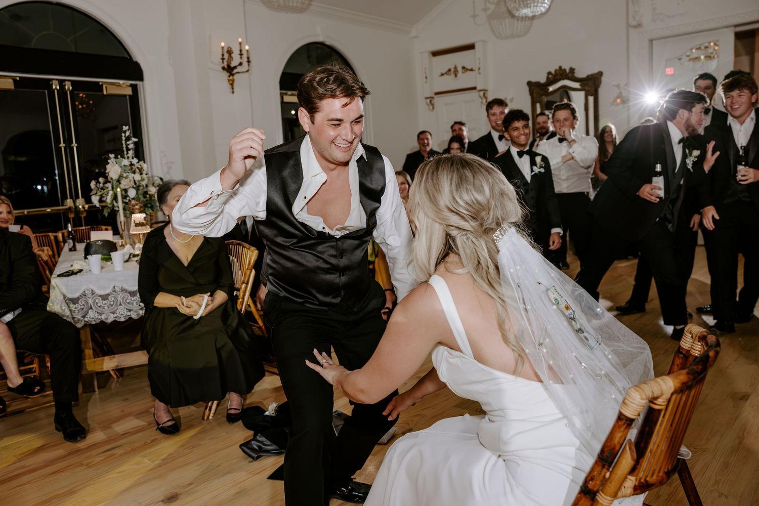 Bride in a white gown and veil dances with a groomsman in a black vest as wedding guests watch in an elegant reception hall.