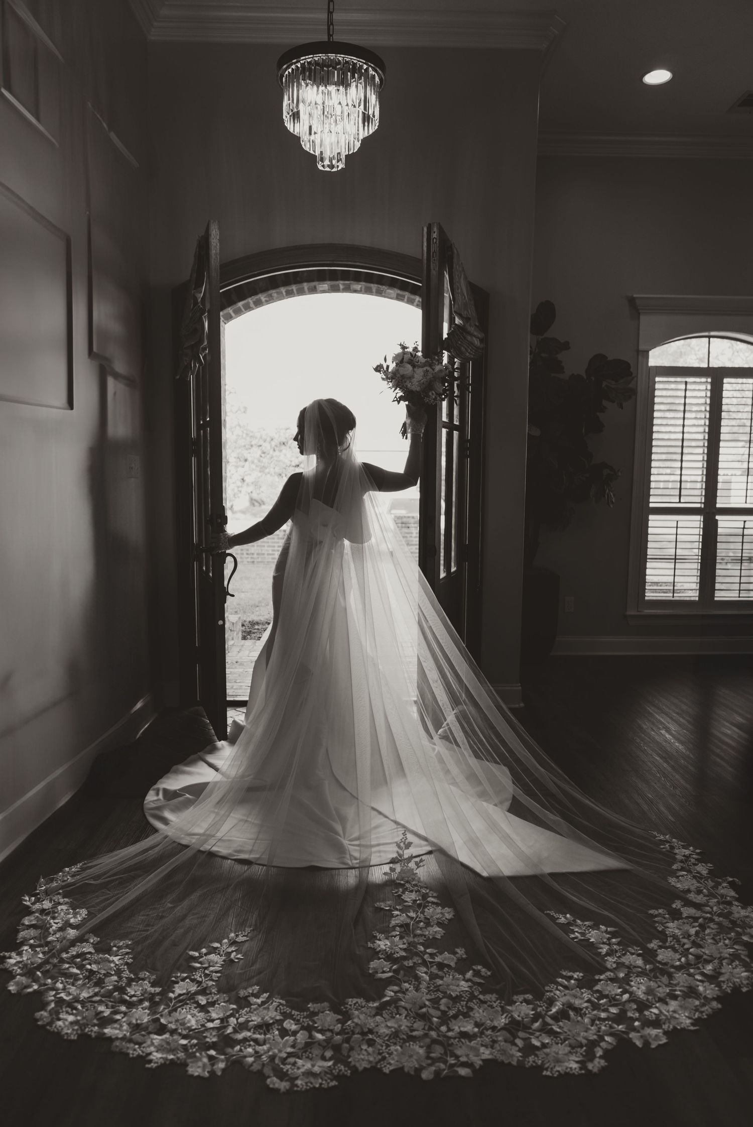 Silhouette of a bride in flowing wedding gown and long veil standing in an elegant doorway with a crystal chandelier above.