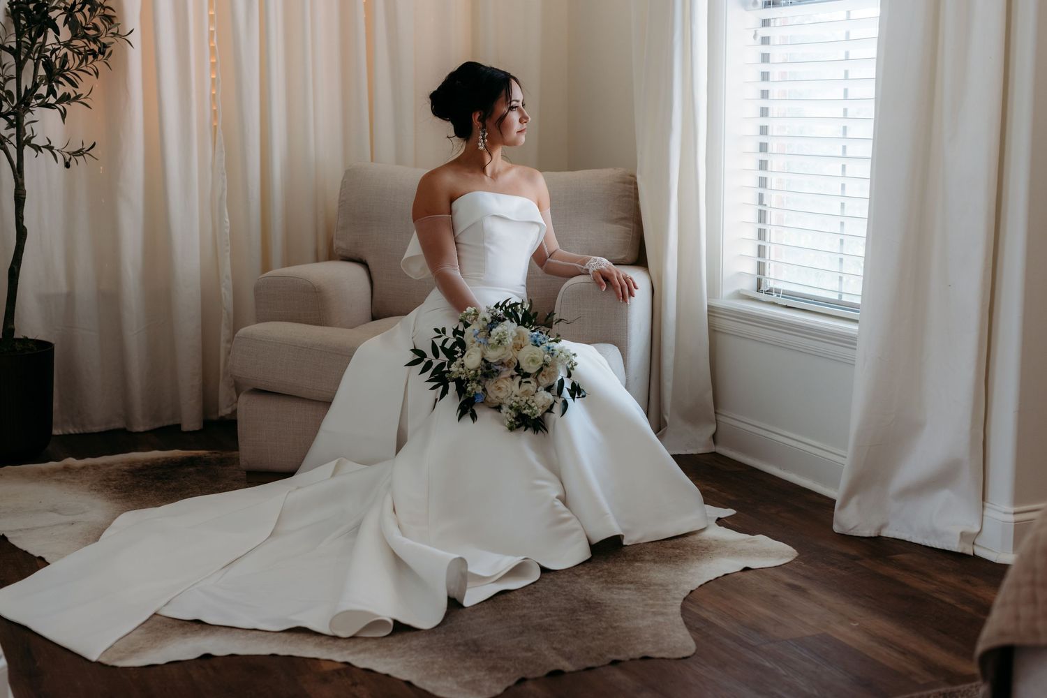 A person in an elegant white wedding gown sits gracefully on a beige couch holding a beautiful floral bouquet.