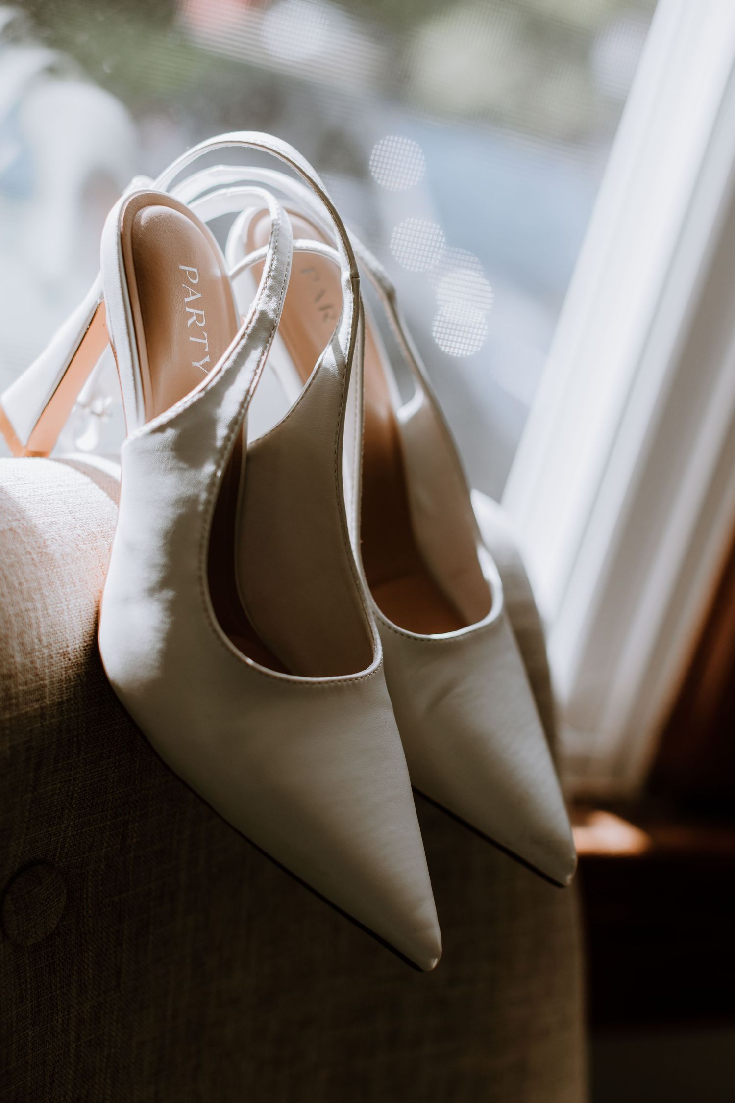 Elegant white slingback pointed-toe heels resting on a chair near a sunlit window.