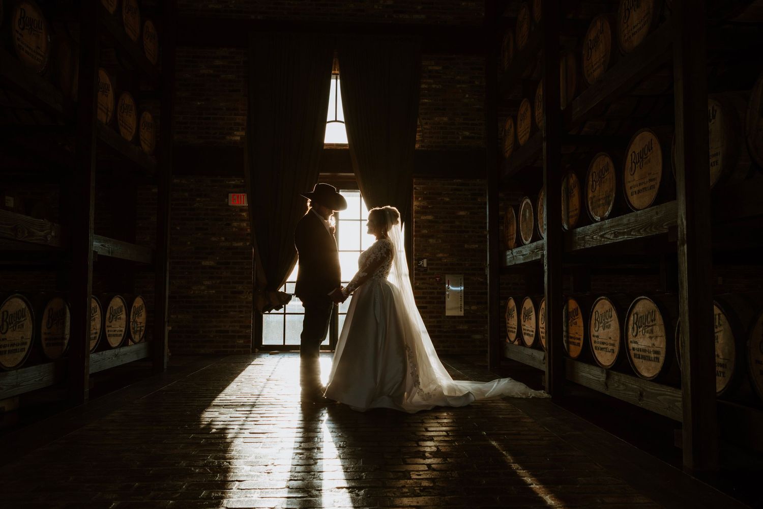 Bride and groom holding hands in a dimly lit bourbon barrel warehouse, backlit by a window.