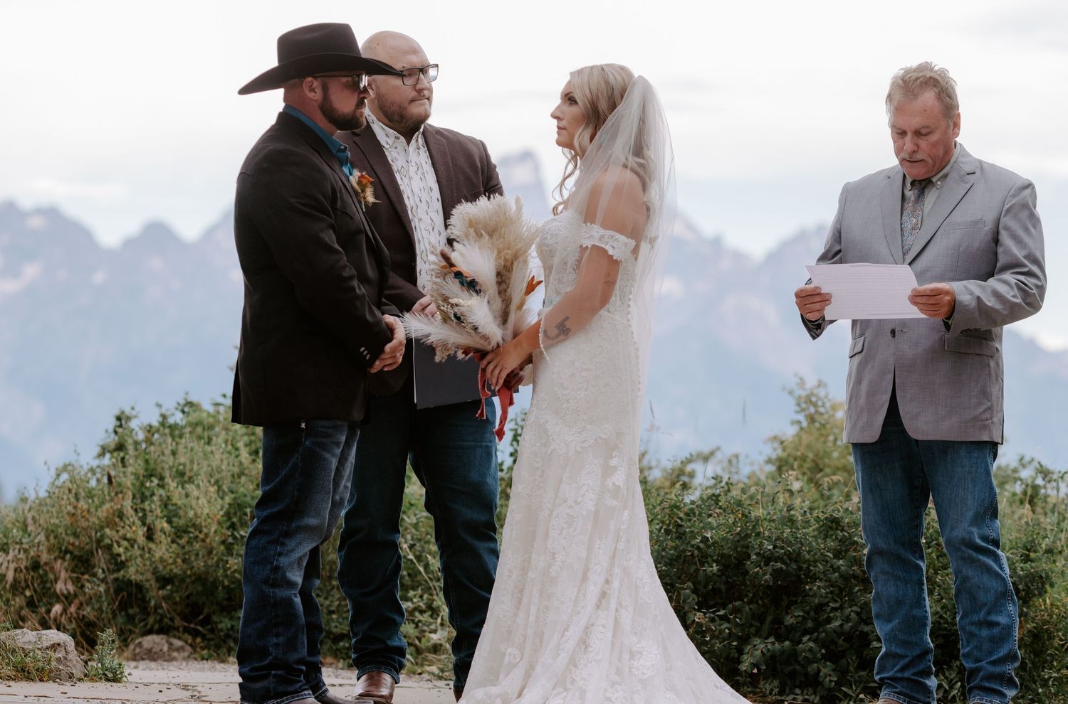 Outdoor mountain wedding ceremony with bride in lace gown, groom in black cowboy hat, officiant reading vows.
