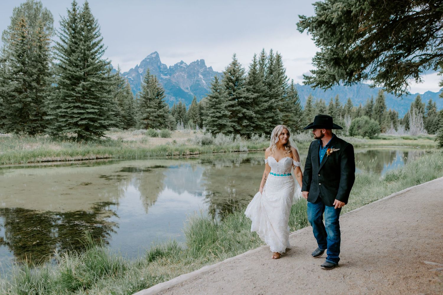 Bride and groom walking hand in hand near a mountain lake with Grand Teton peaks and pine trees in the background.