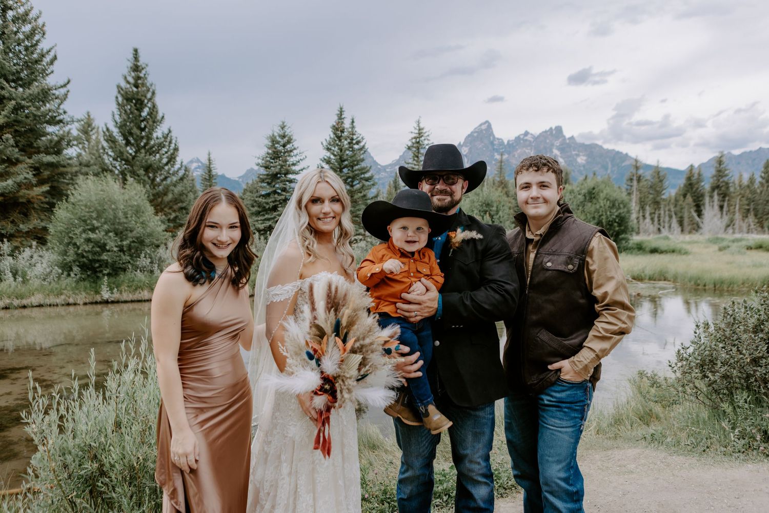 Wedding couple with family posing outdoors near a mountain lake, bride in white gown holding bouquet, groom in black cowboy hat.