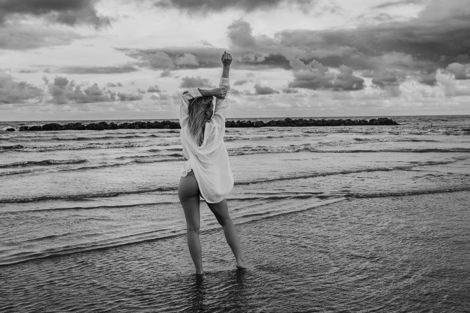 A silhouetted figure in a flowing shirt dress stands in shallow ocean waves at the beach under dramatic clouds.