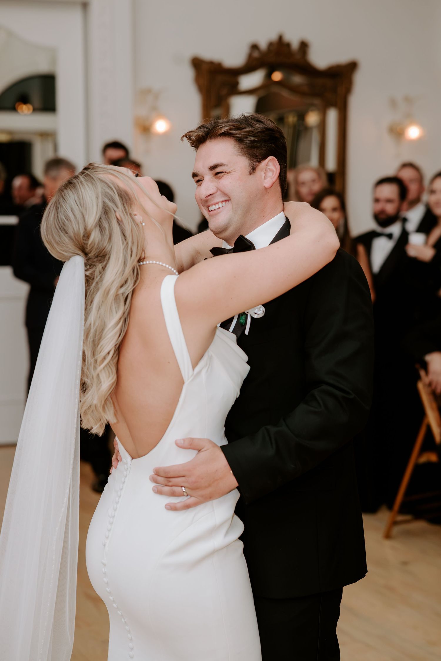 Smiling groom in a black tuxedo embraces a bride in a white open-back gown and veil during their first dance as wedding guests watch.