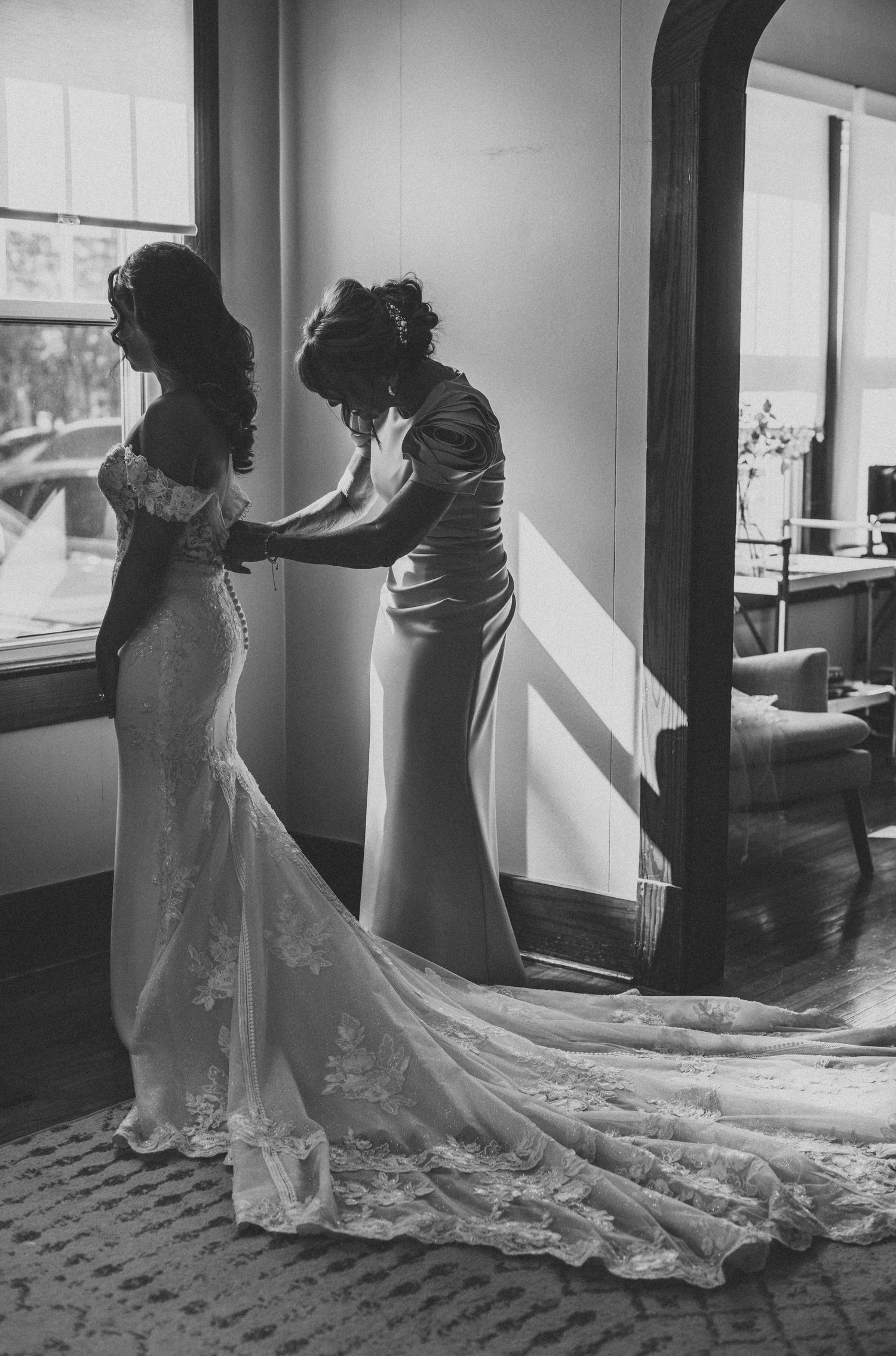 Black and white photo of a bride in a lace gown being helped by a bridesmaid before her wedding ceremony.
