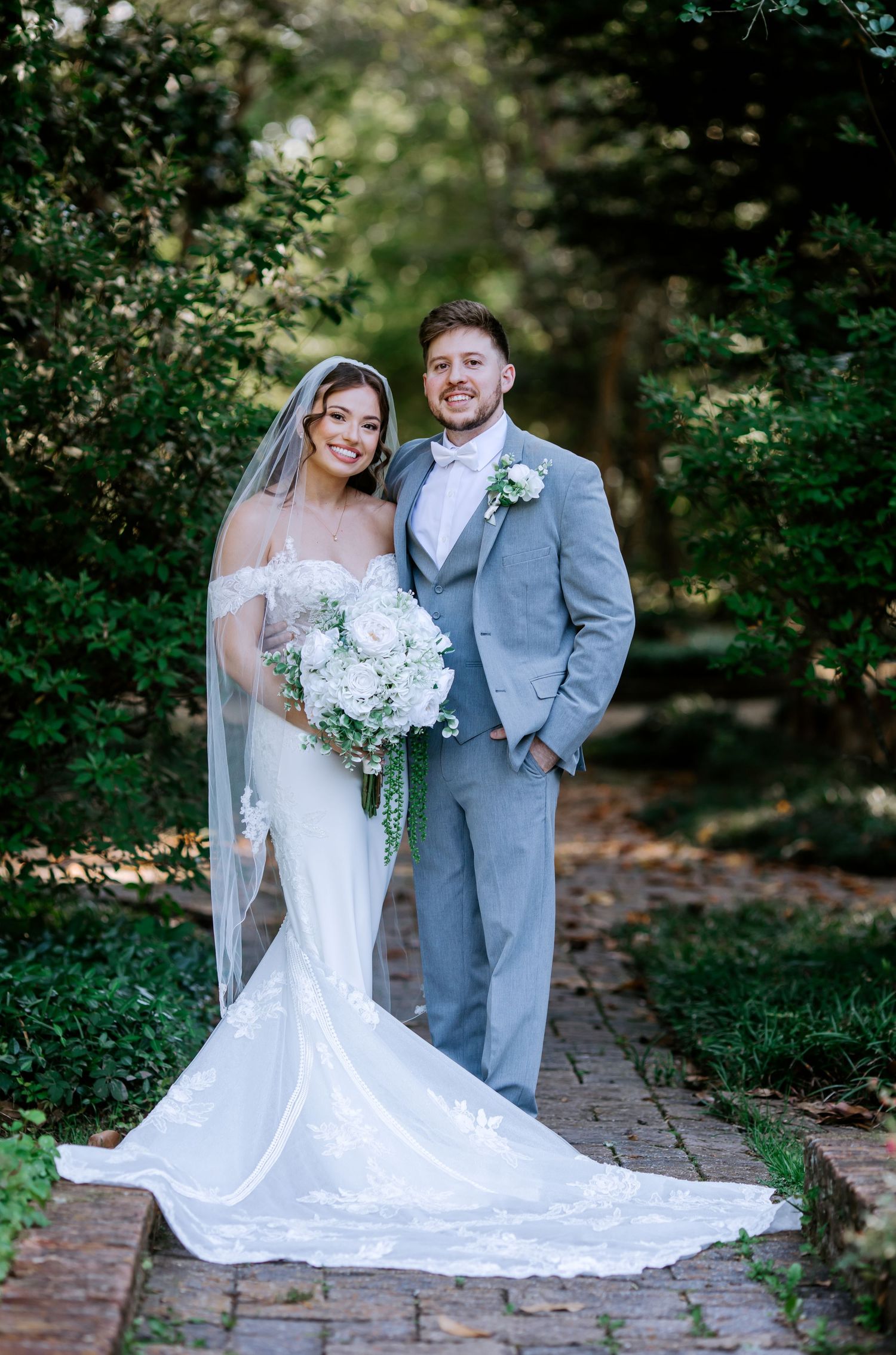 Smiling bride and groom posing together outdoors, bride in lace gown with long veil, groom in light gray suit.