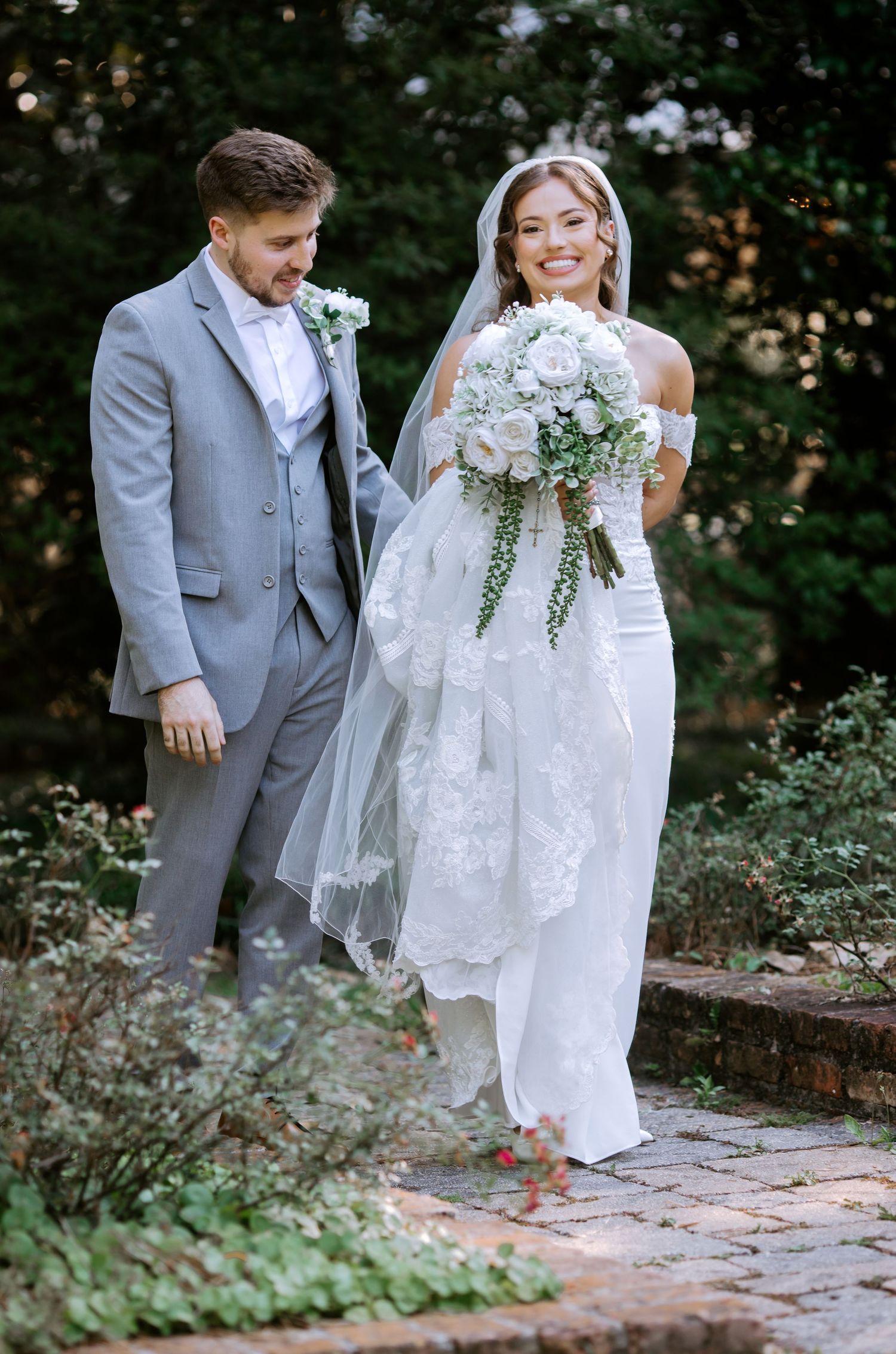 Smiling bride in off-shoulder lace gown holding white bouquet walks with groom in gray suit on garden path.