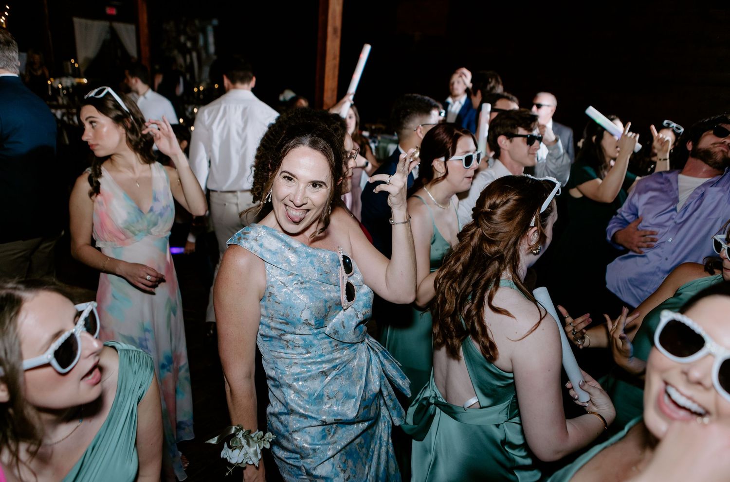 Bride and bridesmaids in teal dresses dancing with glow sticks and white sunglasses at wedding reception.